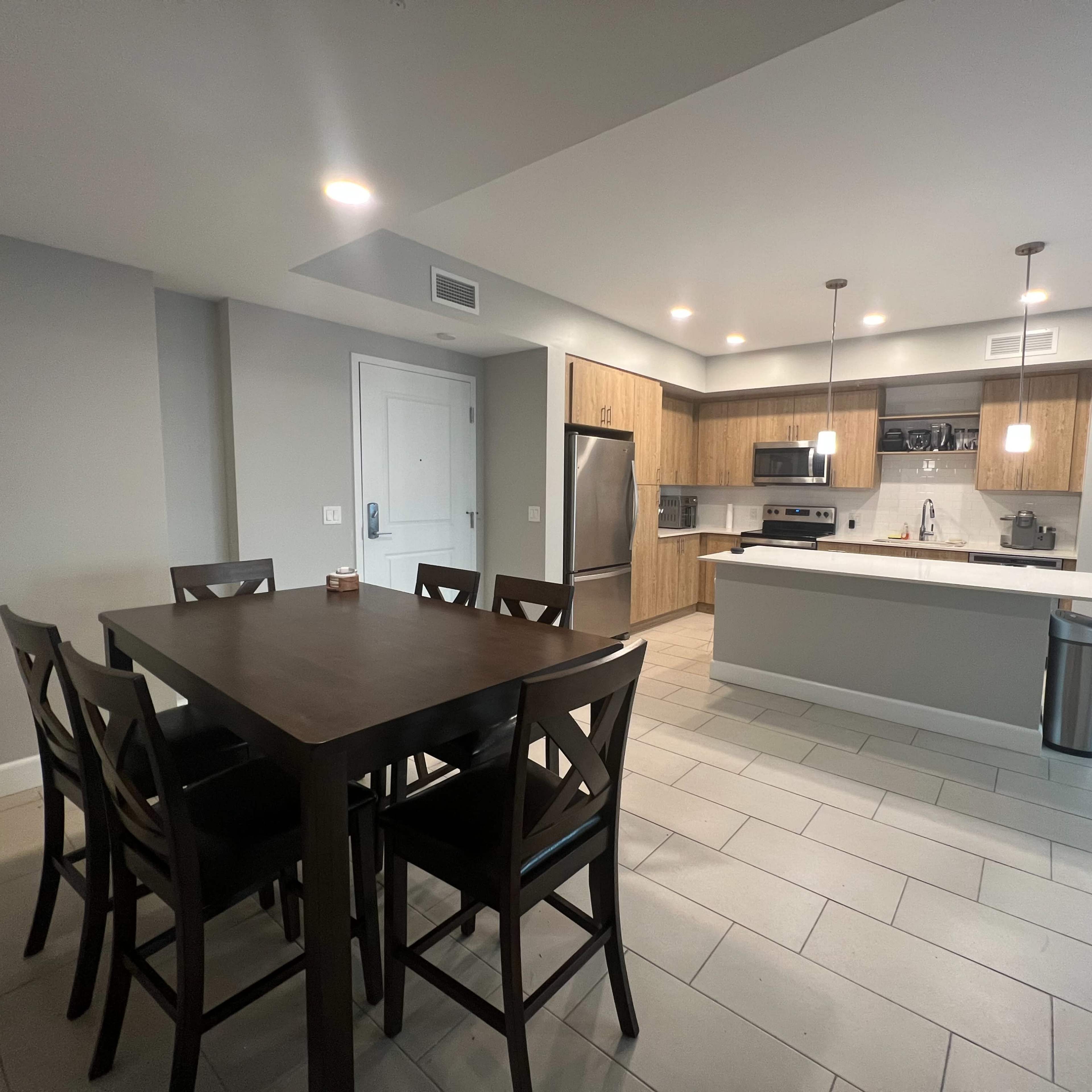 A modern kitchen and dining area with a wooden dining table, black chairs, and a kitchen featuring light wood cabinetry and stainless steel appliances.