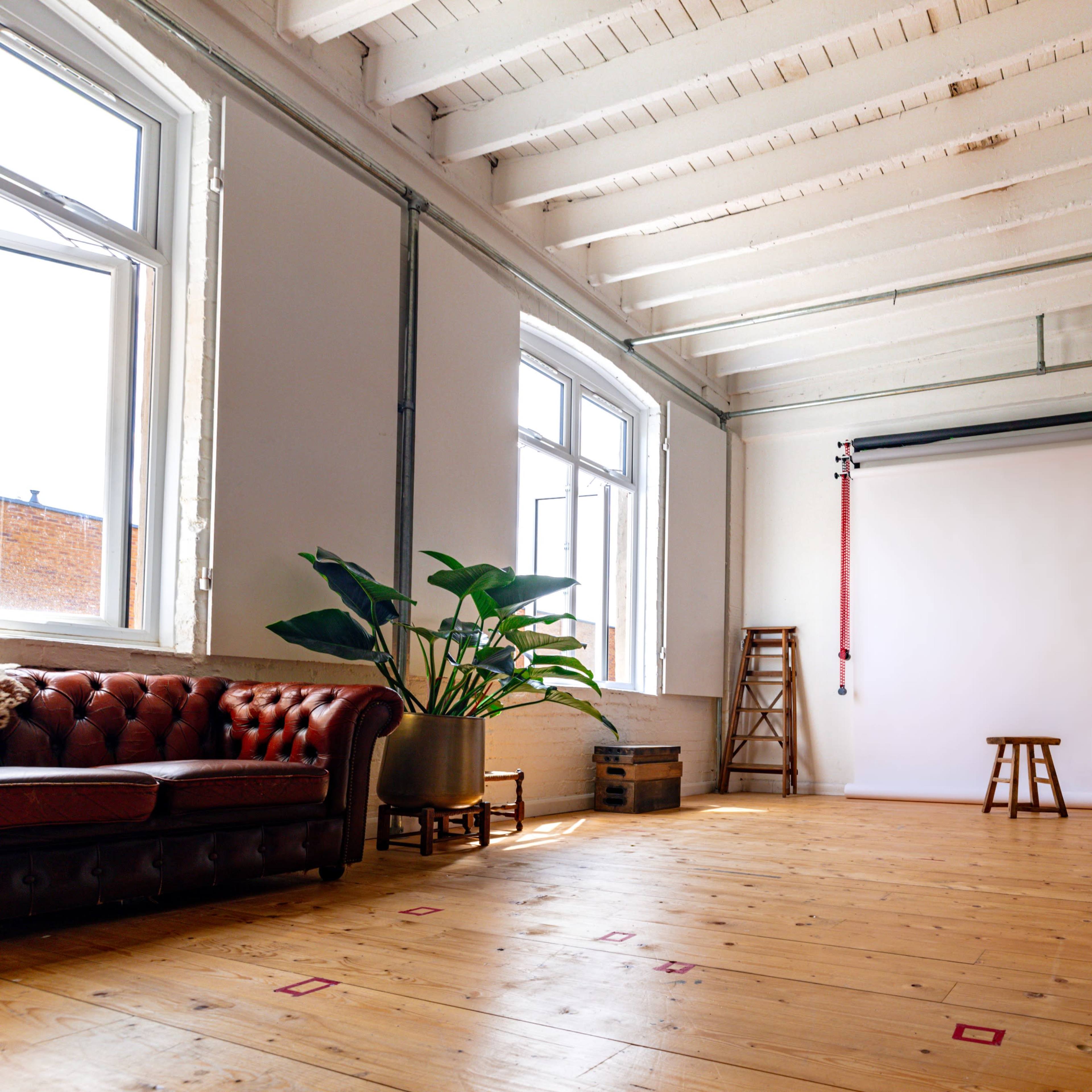 A spacious studio with high ceilings, a large white backdrop, a wooden floor, a brown leather couch, a stool, and a potted plant.
