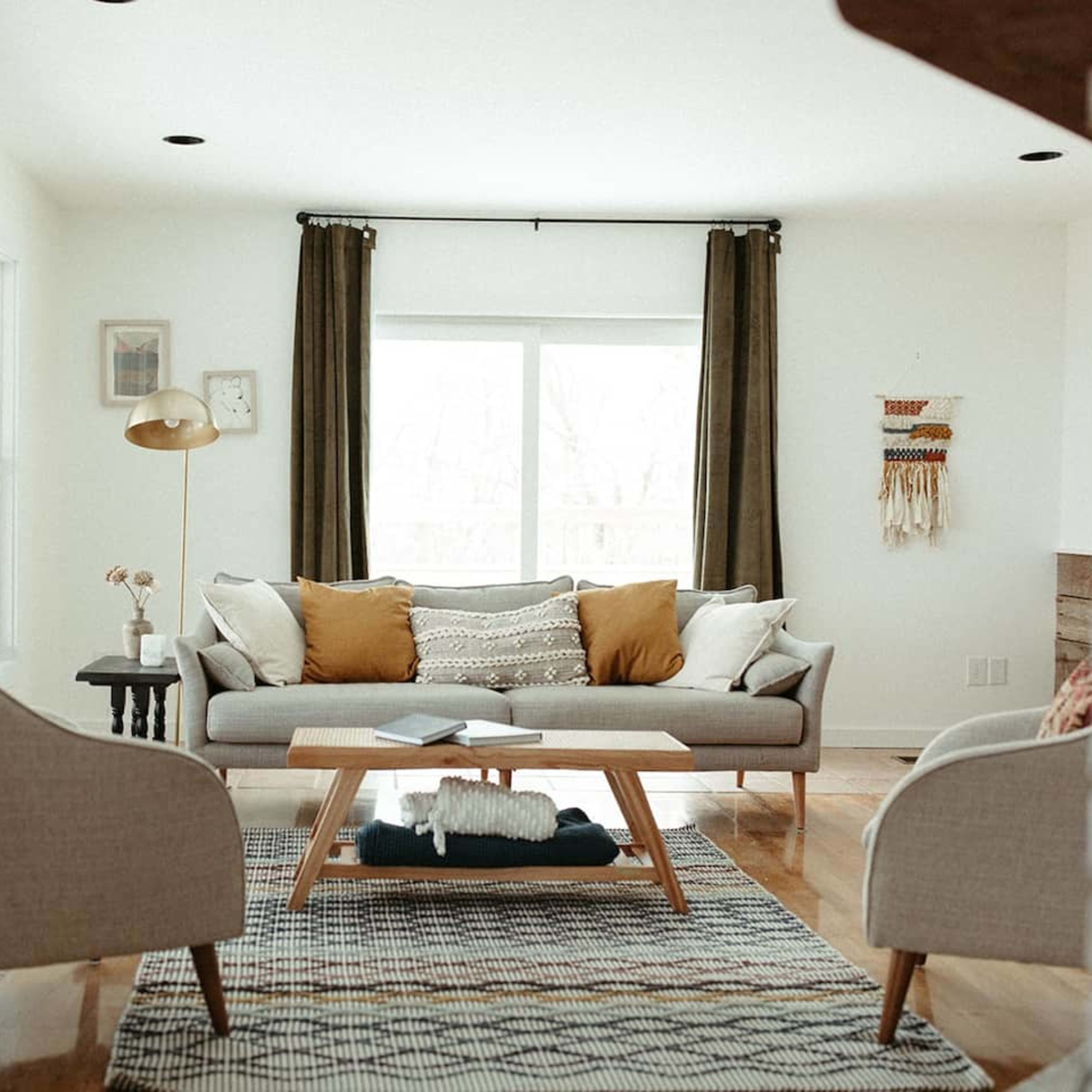 A modern living room features a gray sofa with decorative pillows, two armchairs, a coffee table, and large windows with curtains.