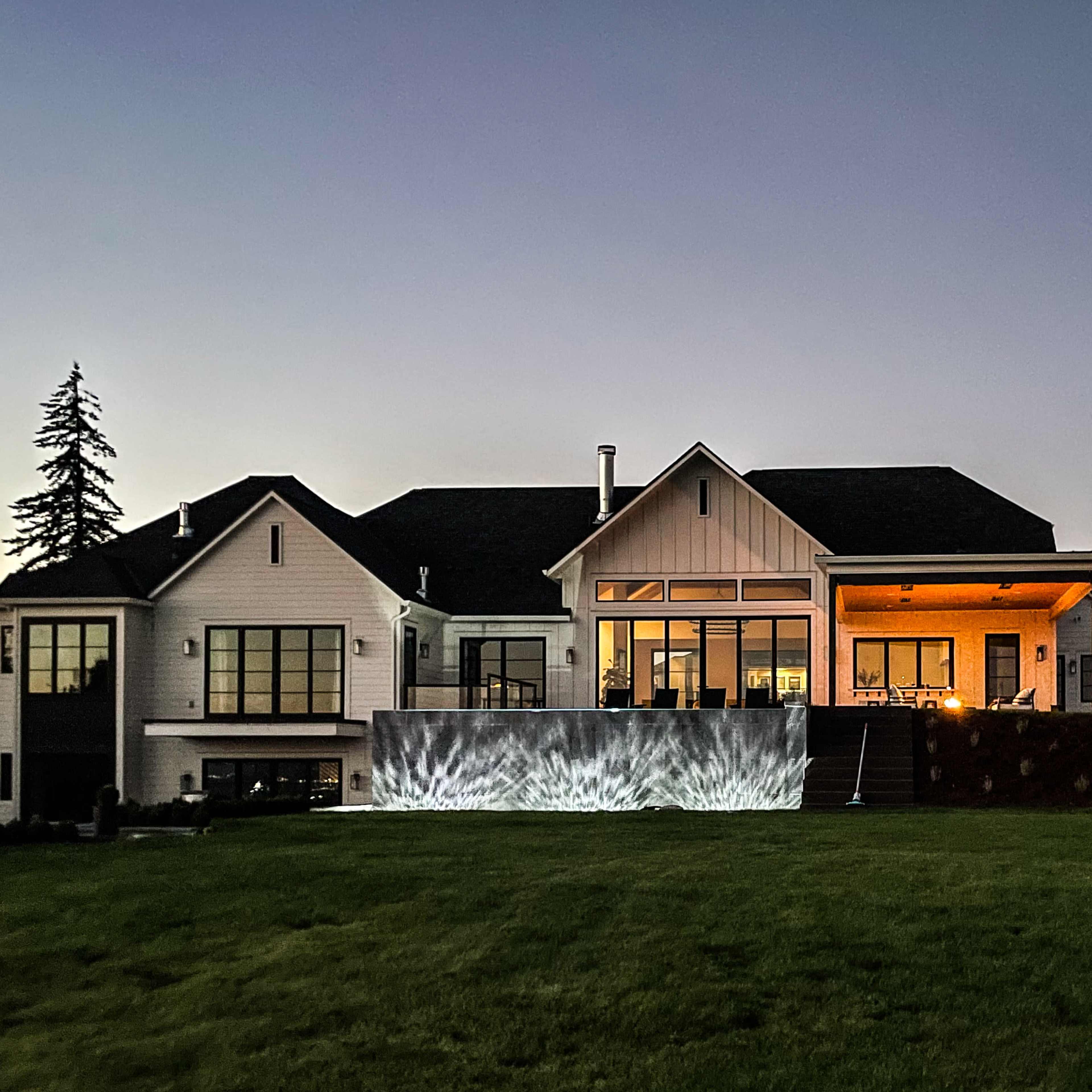 A modern house with large windows and a deck features a light display on the patio, set against a twilight sky.