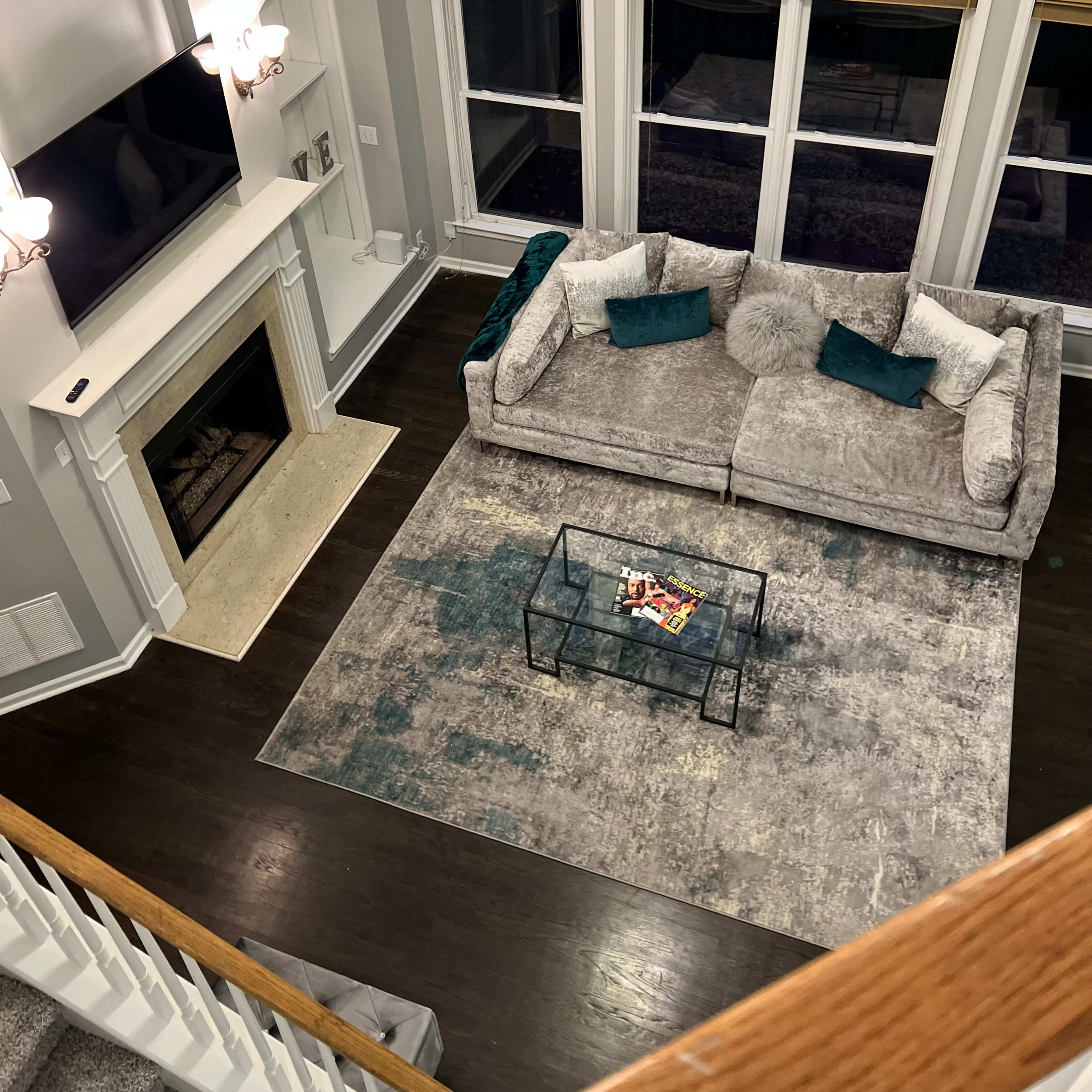 A spacious living room features a large gray couch, a glass coffee table, and a modern fireplace beneath a wall-mounted television, surrounded by tall windows.