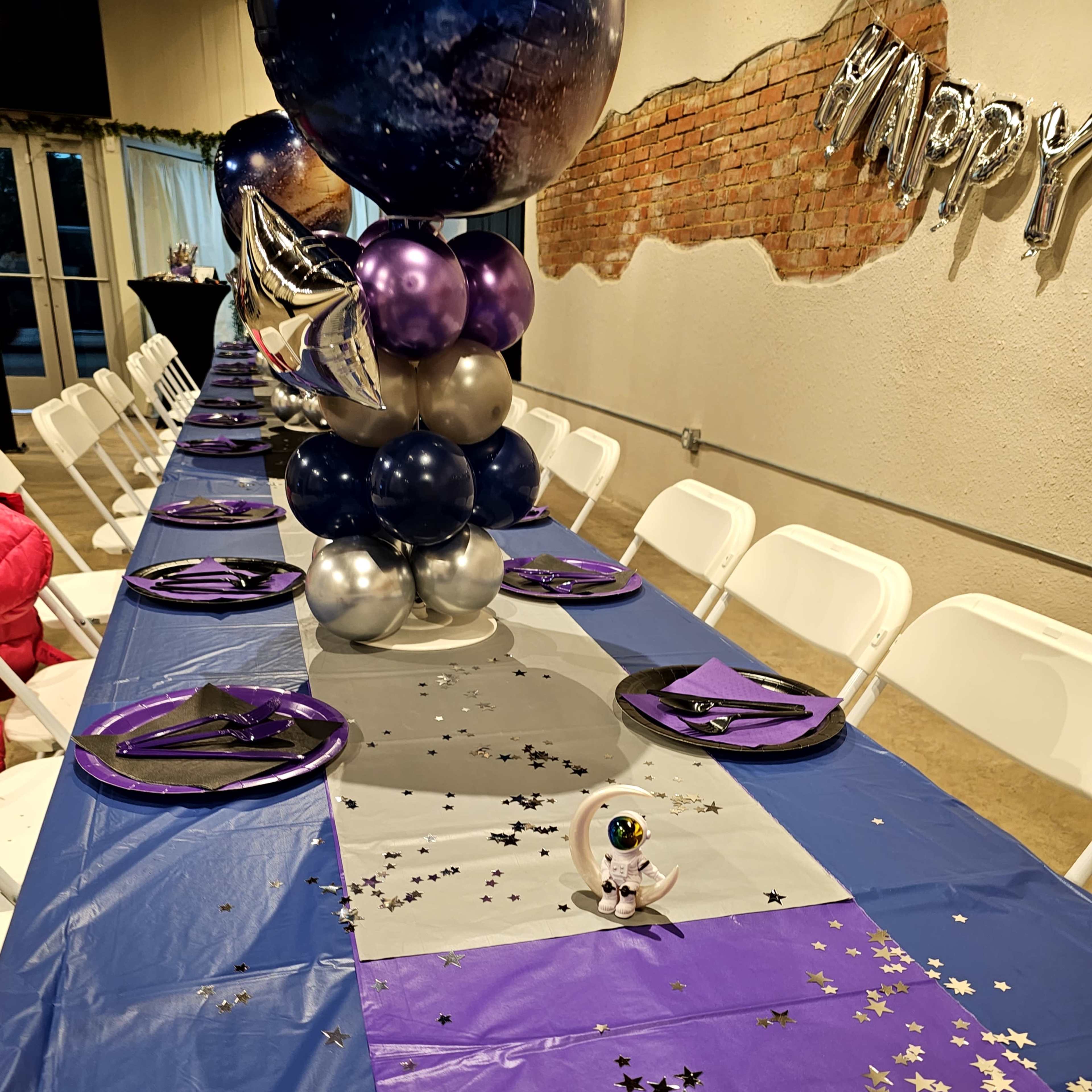 A long table decorated with purple and silver tablecloths, balloons, plates, and star confetti is set for a celebration.