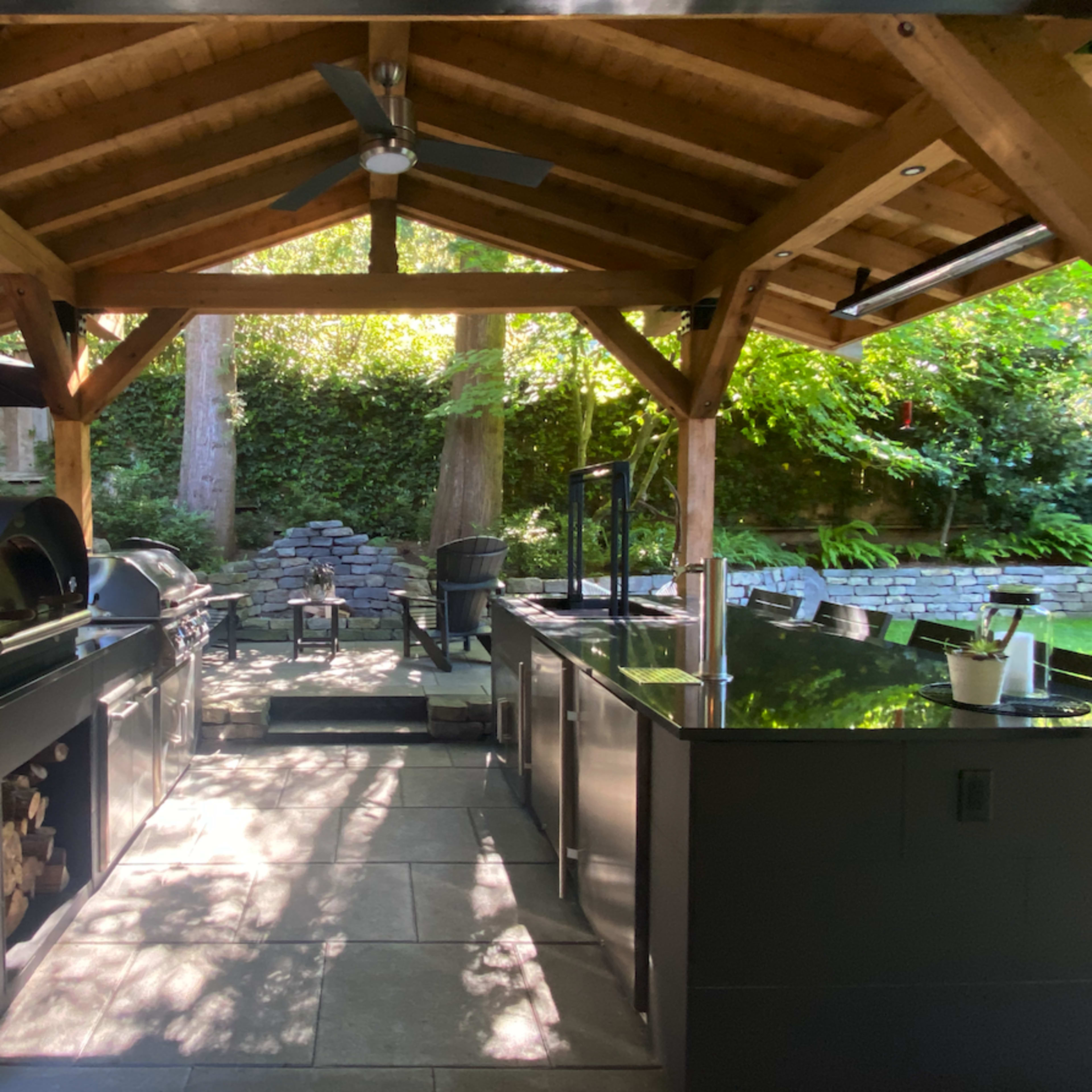 An outdoor kitchen under a wooden structure, equipped with a grill, pizza oven, and counter space, surrounded by greenery.