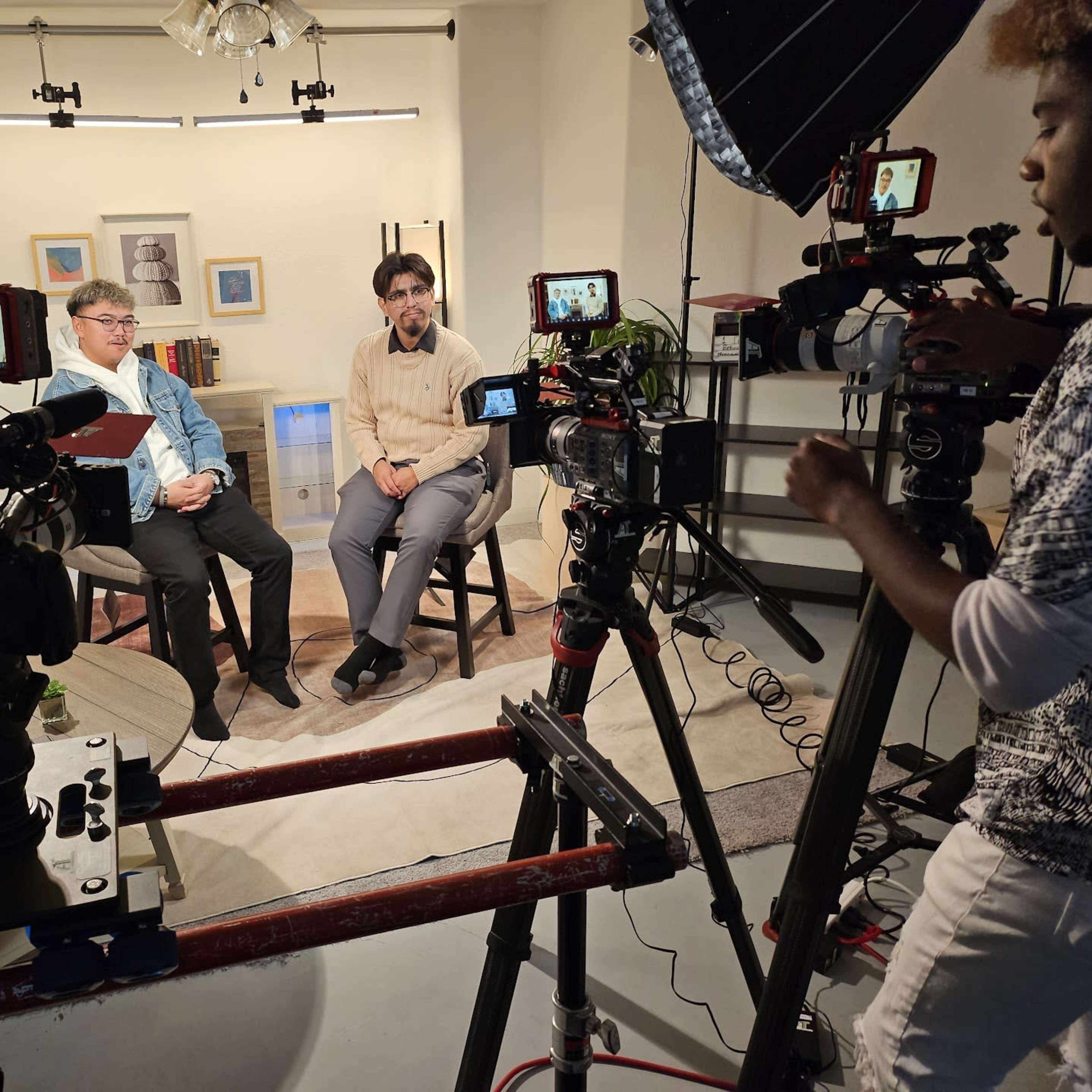 Two men sit in chairs for an interview, surrounded by multiple cameras and lighting equipment in a studio setting.