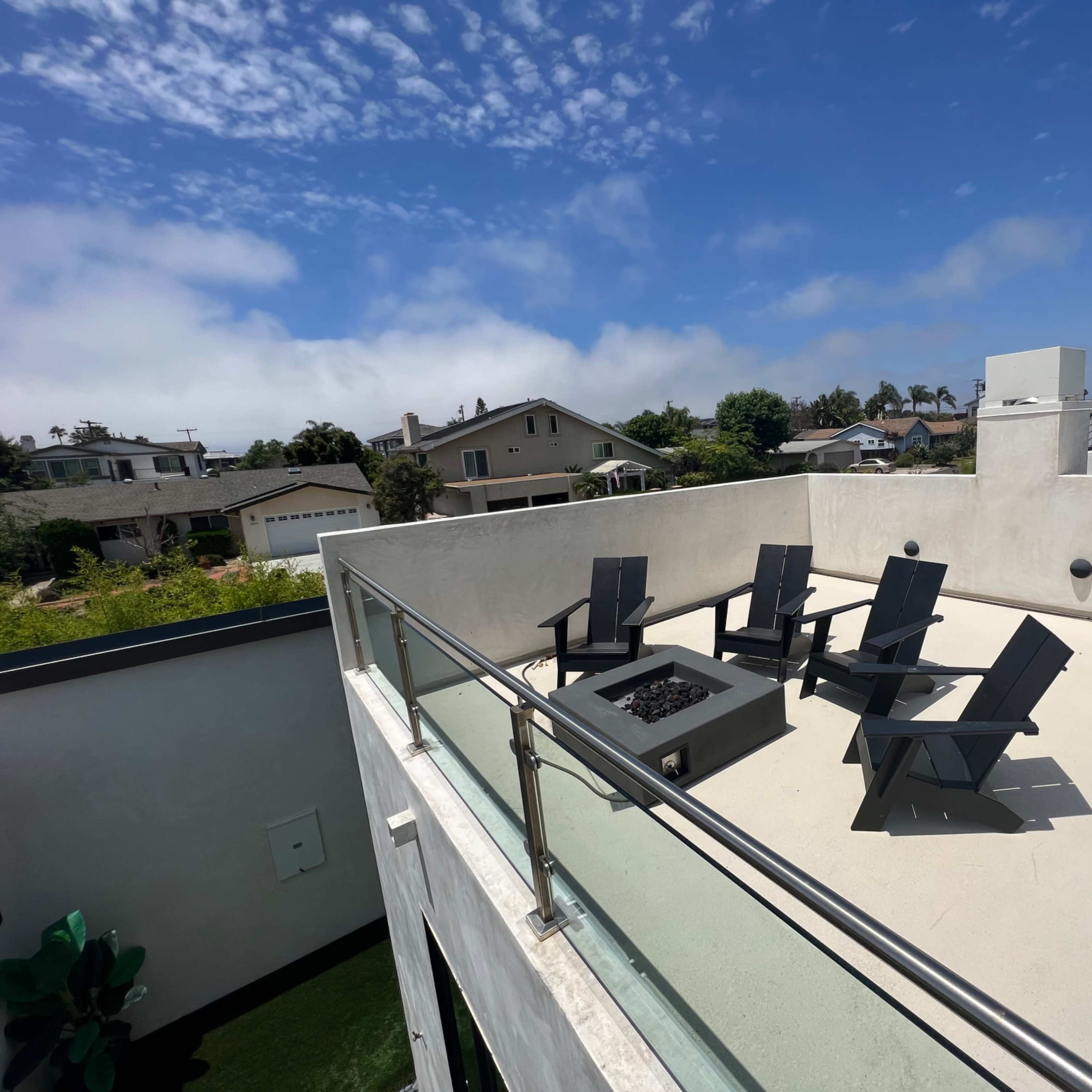 An outdoor rooftop area featuring a fire pit surrounded by five black chairs under a partly cloudy sky.