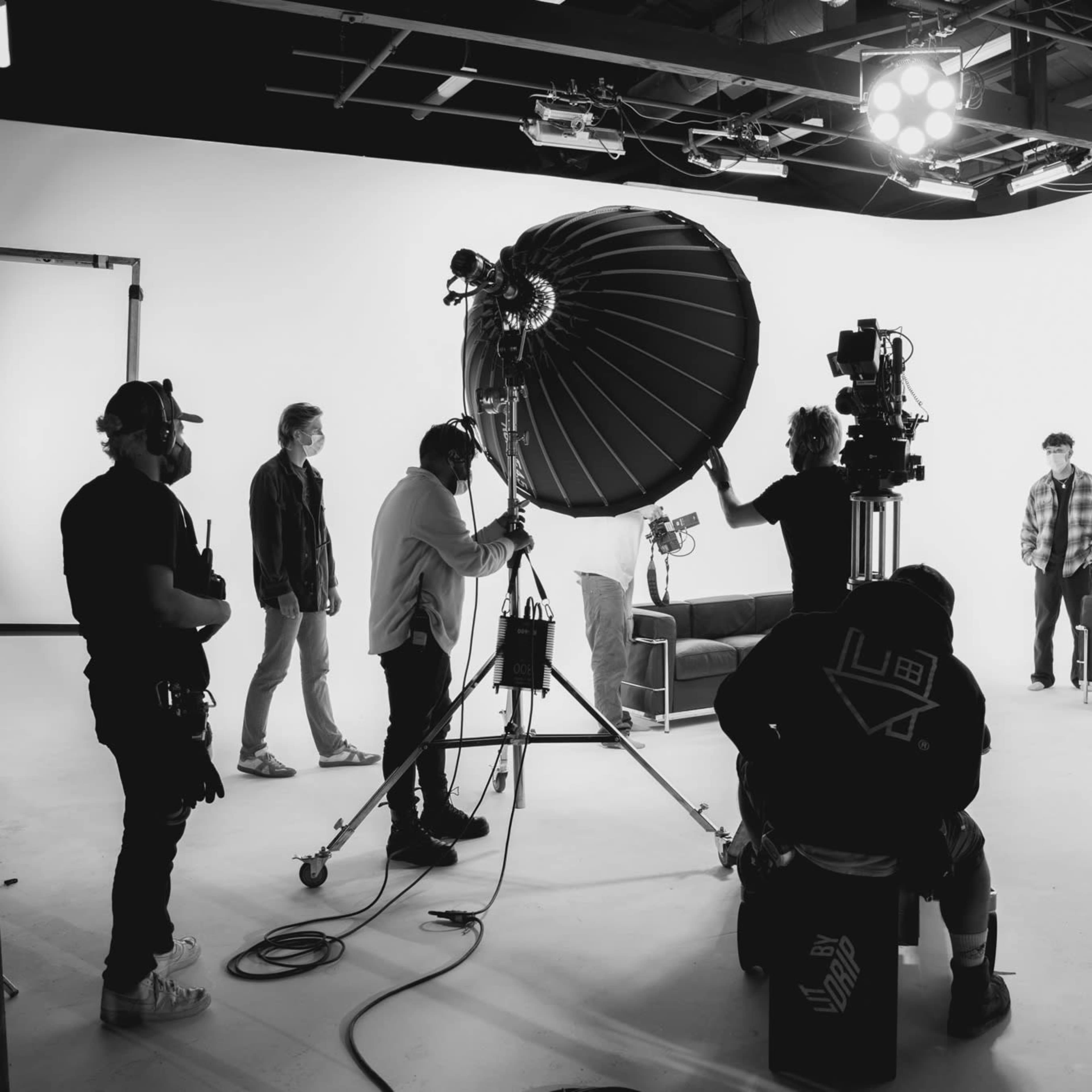 A group of crew members operates equipment on a film set while two individuals stand in front of a backdrop near two couches.