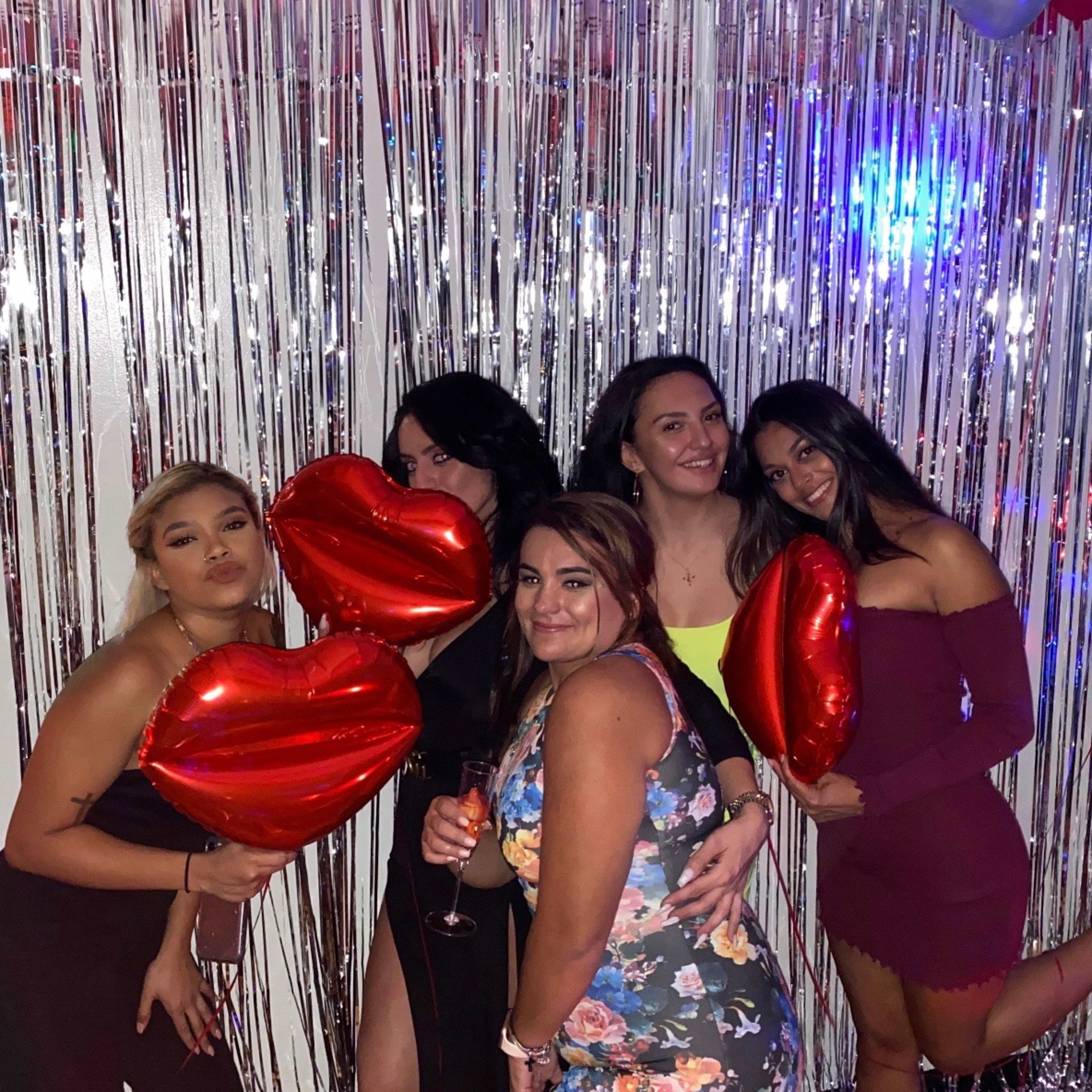 A group of five women poses with large red heart-shaped balloons in front of a shiny silver backdrop.