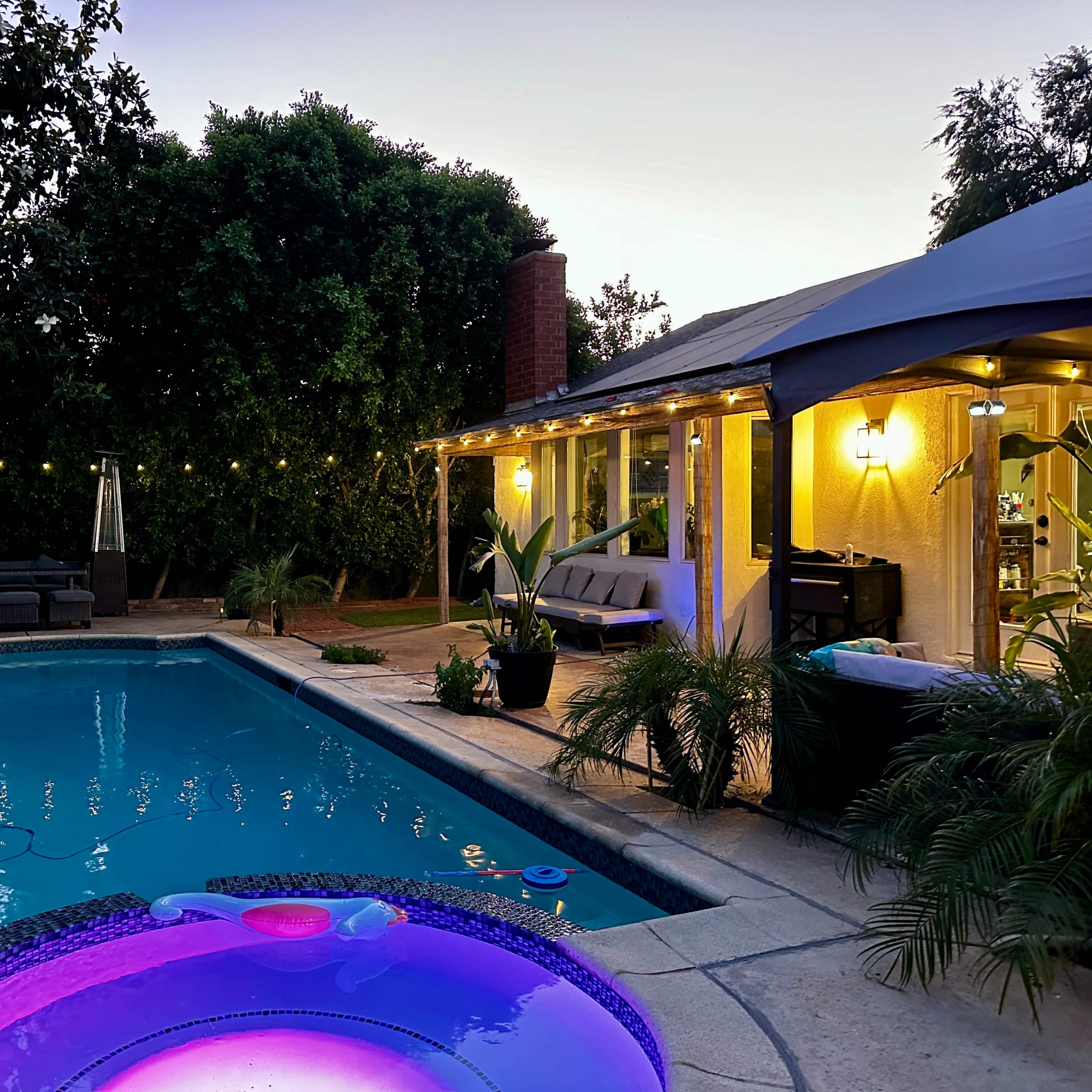 The scene depicts a backyard with a swimming pool, a hot tub illuminated in purple, and a cozy seating area, all surrounded by lush greenery and soft outdoor lighting as the sun sets.