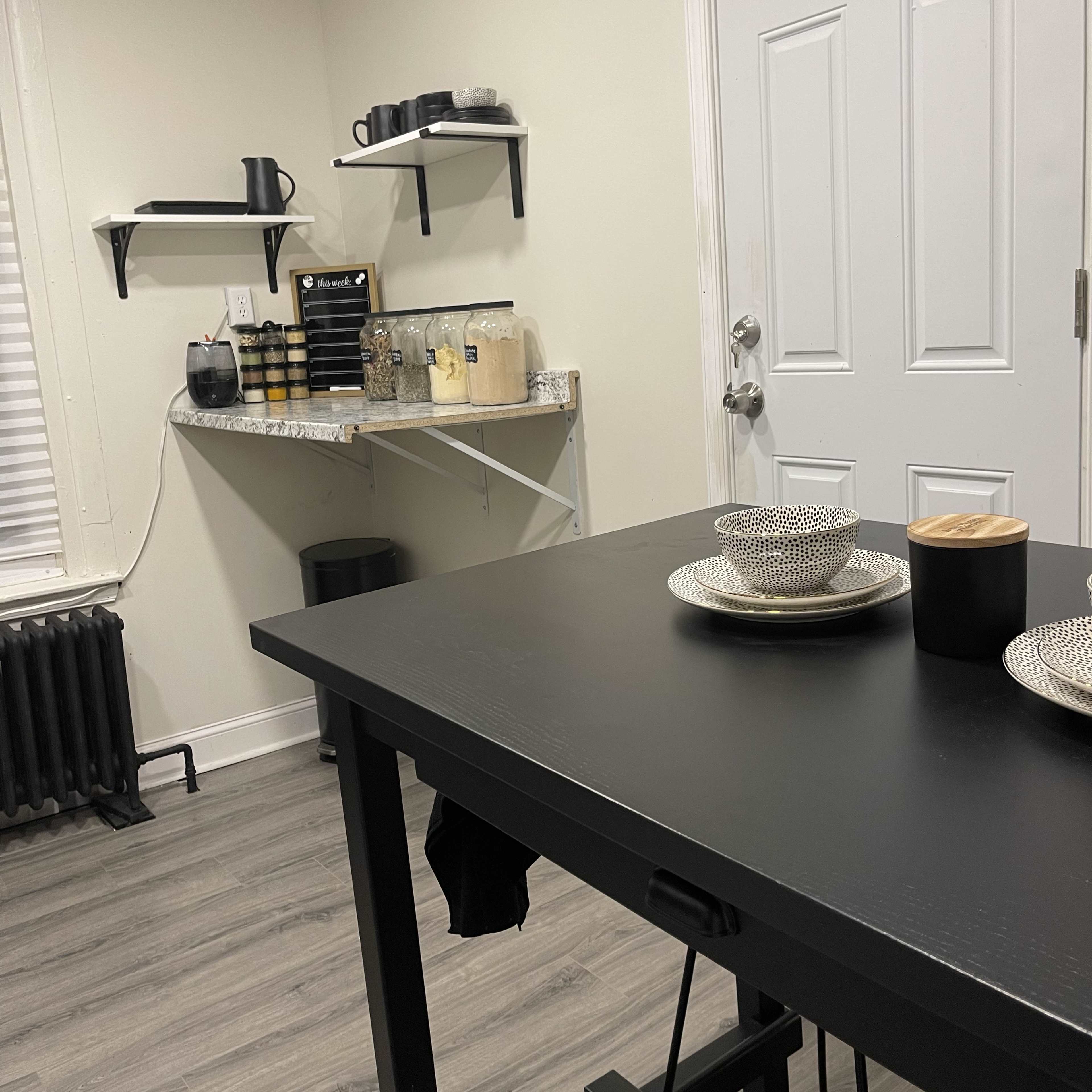 A small kitchen area with a black table set for a meal, featuring stacks of plates and two cups, alongside shelves holding kitchen appliances and utensils.