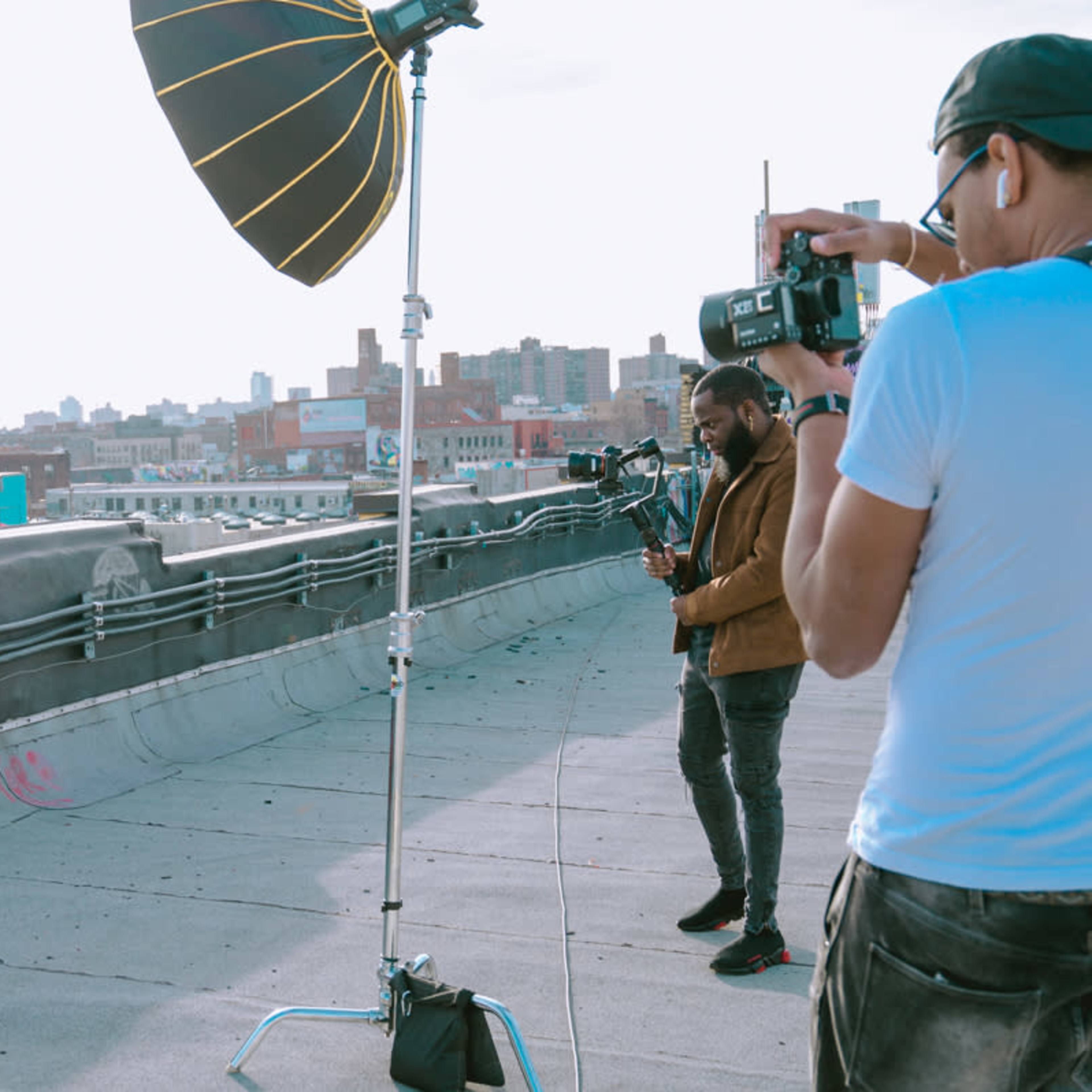 A model poses in a fur coat on a rooftop while a photographer captures the shot with a large reflector in the background.