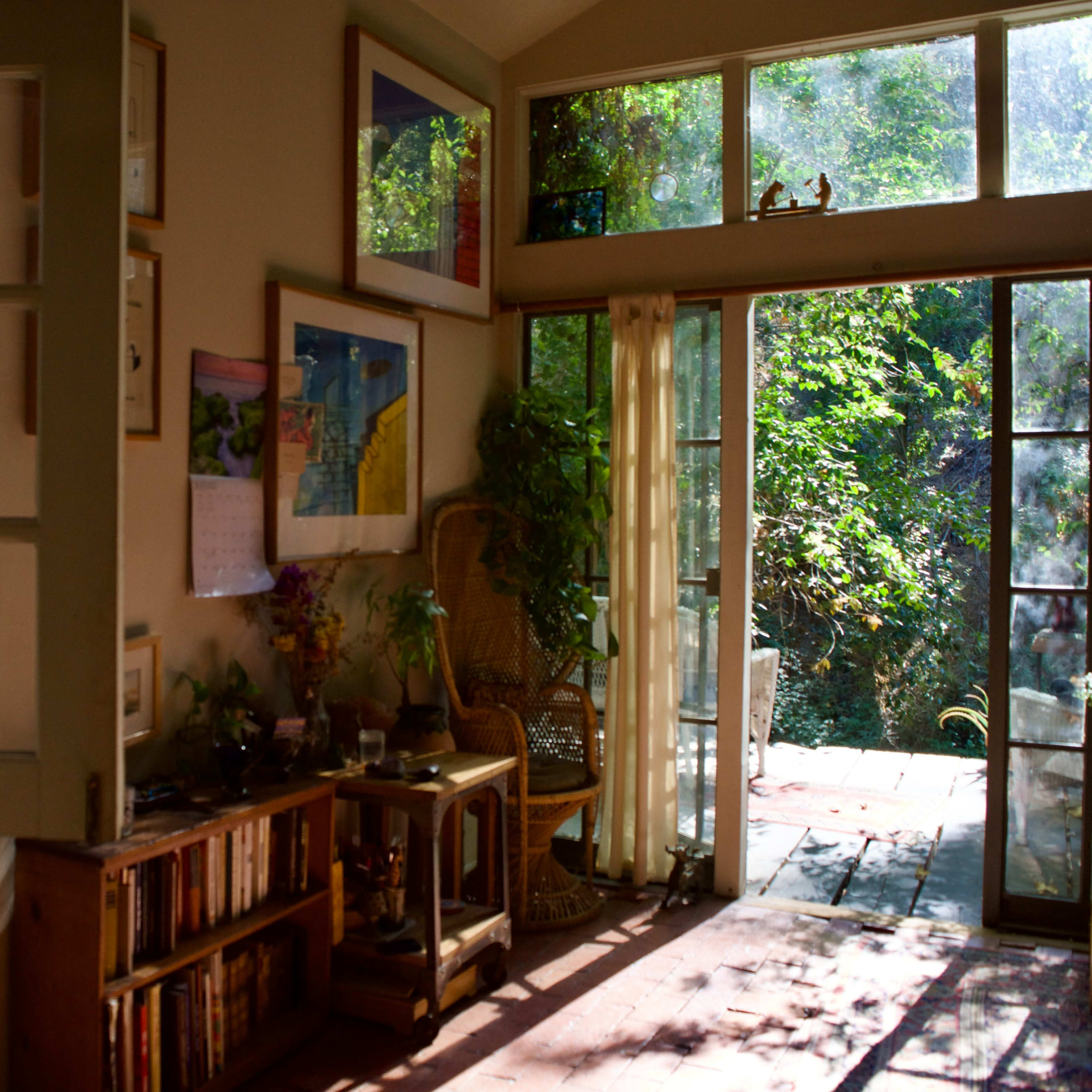 A sunlit room opens to a garden through large glass doors, featuring a wooden table, bookshelves, and framed artwork on the walls.