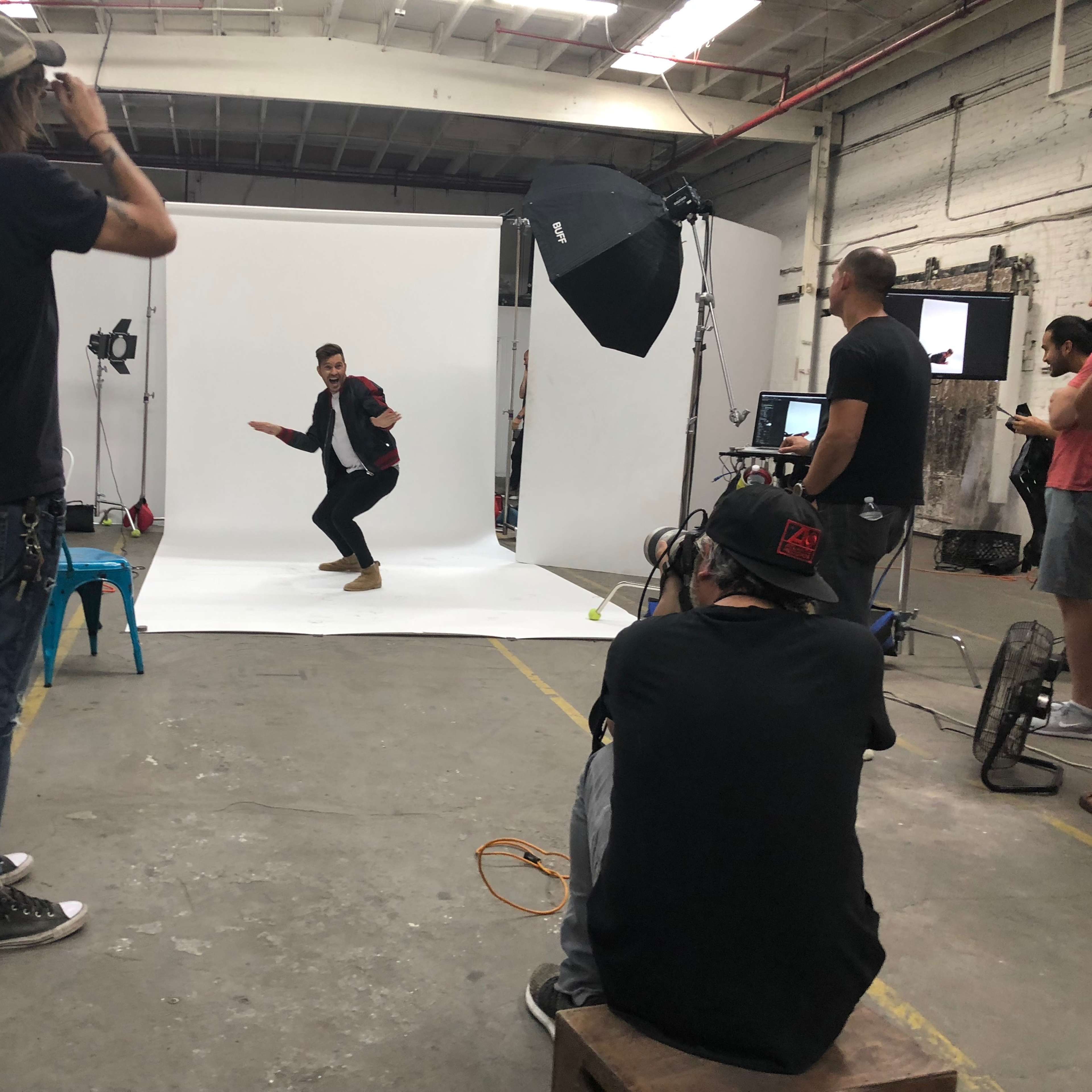 A group of people is working in a photo studio where a man is posing on a white backdrop while others are capturing images and setting up equipment.