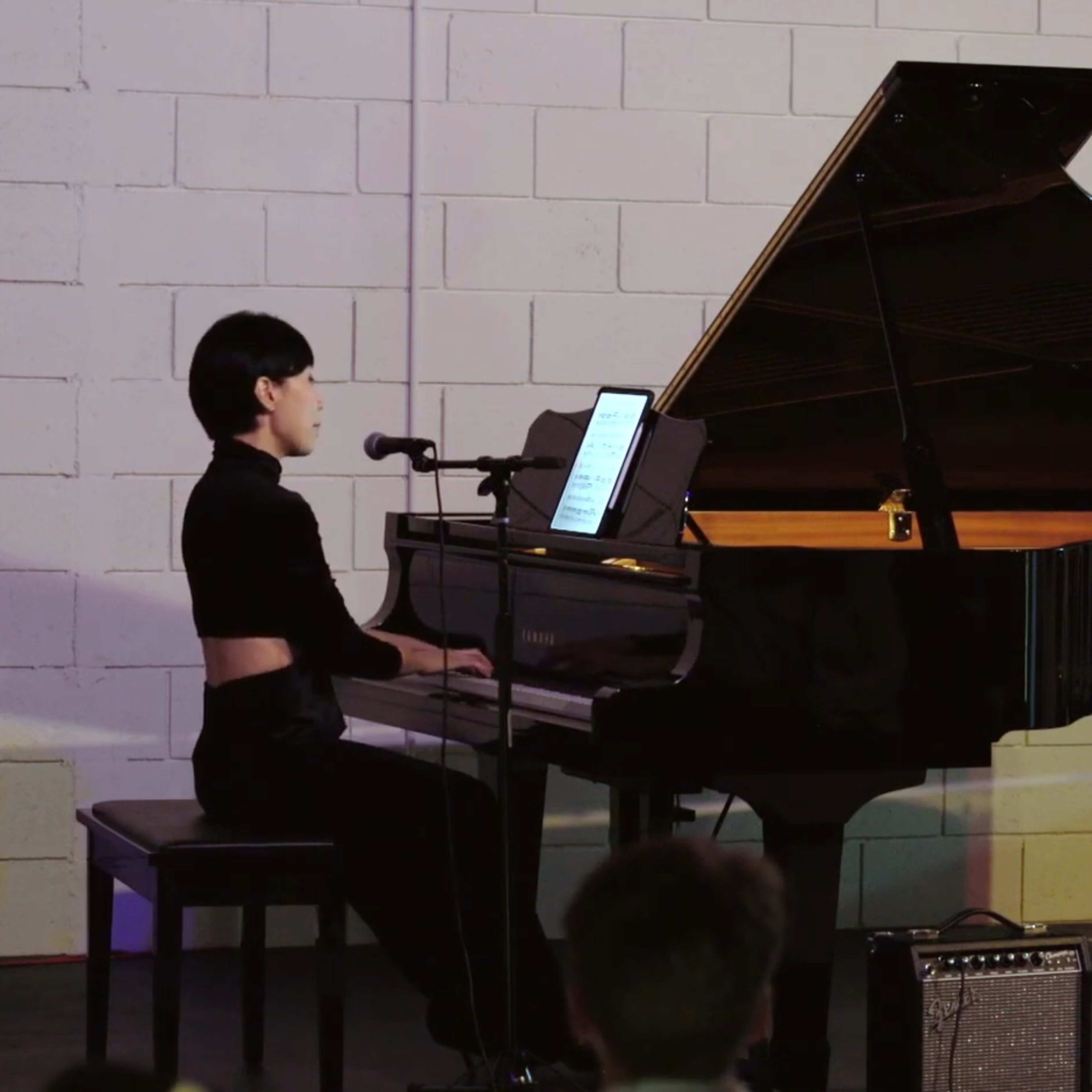A musician plays a grand piano while singing into a microphone in a staged performance setting.