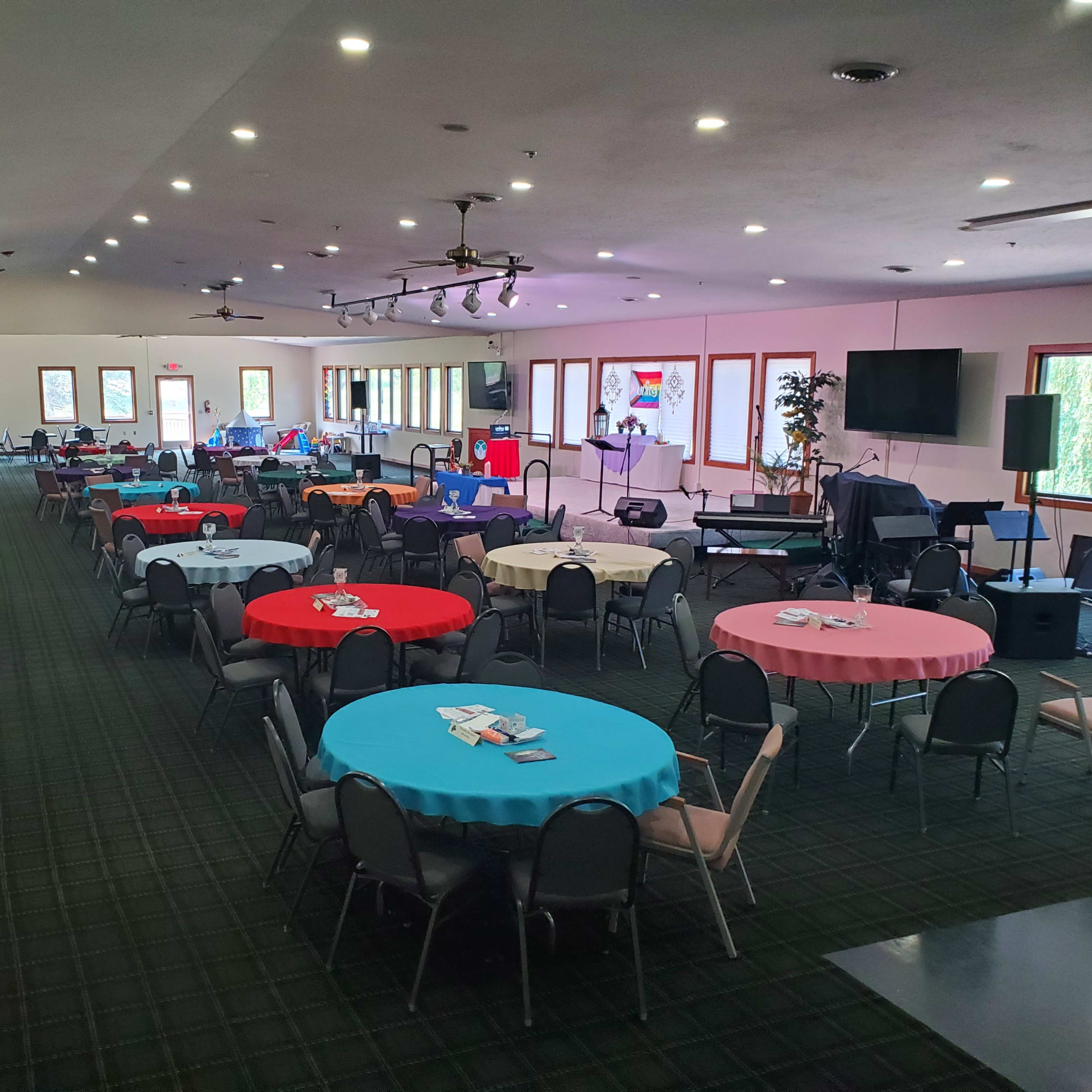 A spacious event hall with several round tables covered in colorful tablecloths, arranged in a well-lit setting with a stage and equipment at one end.