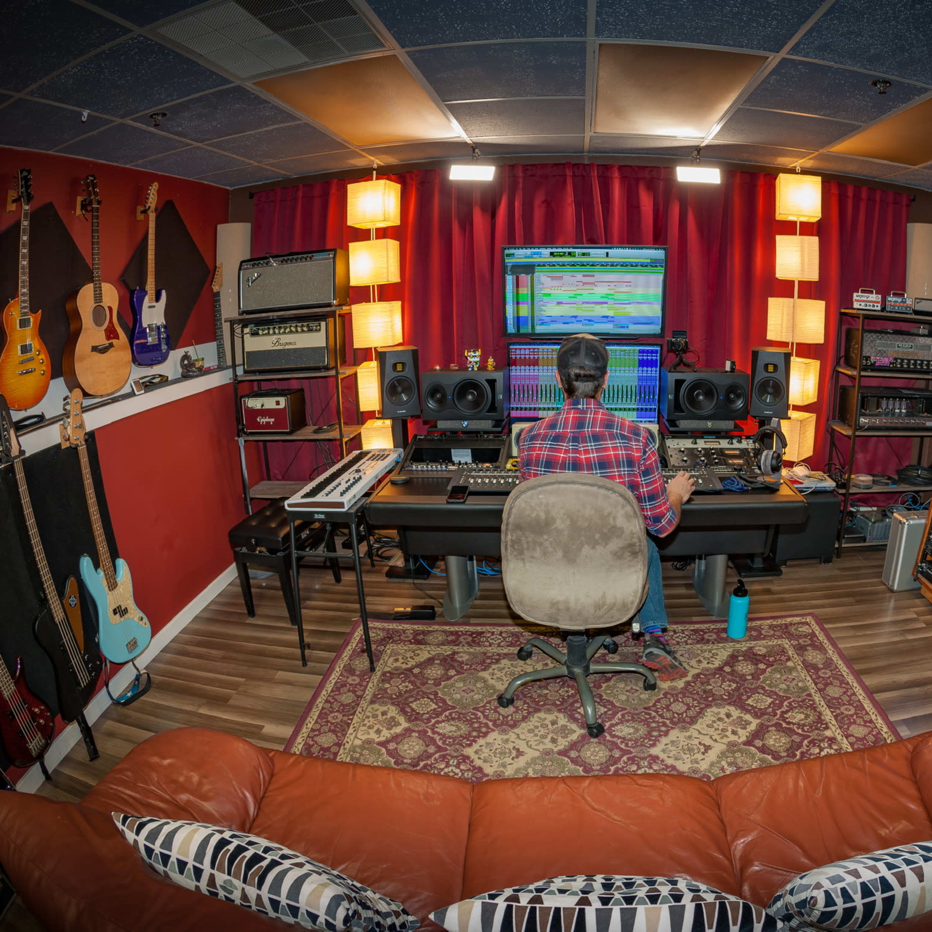 A person sits at a computer workstation in a music studio surrounded by various musical instruments and equipment.