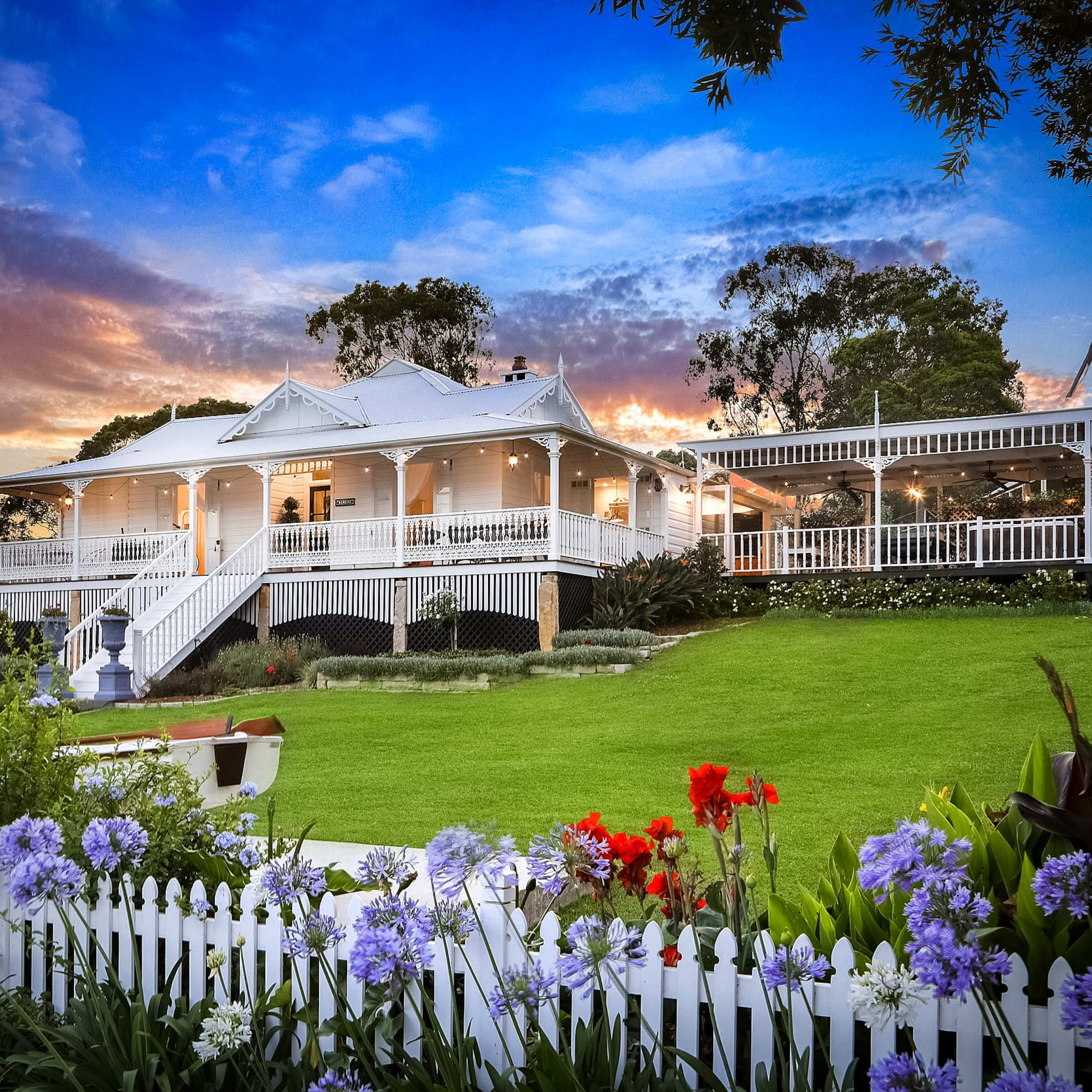 A large, two-story house with a wraparound veranda is set against a colorful sunset, surrounded by well-maintained gardens and vibrant flowers.