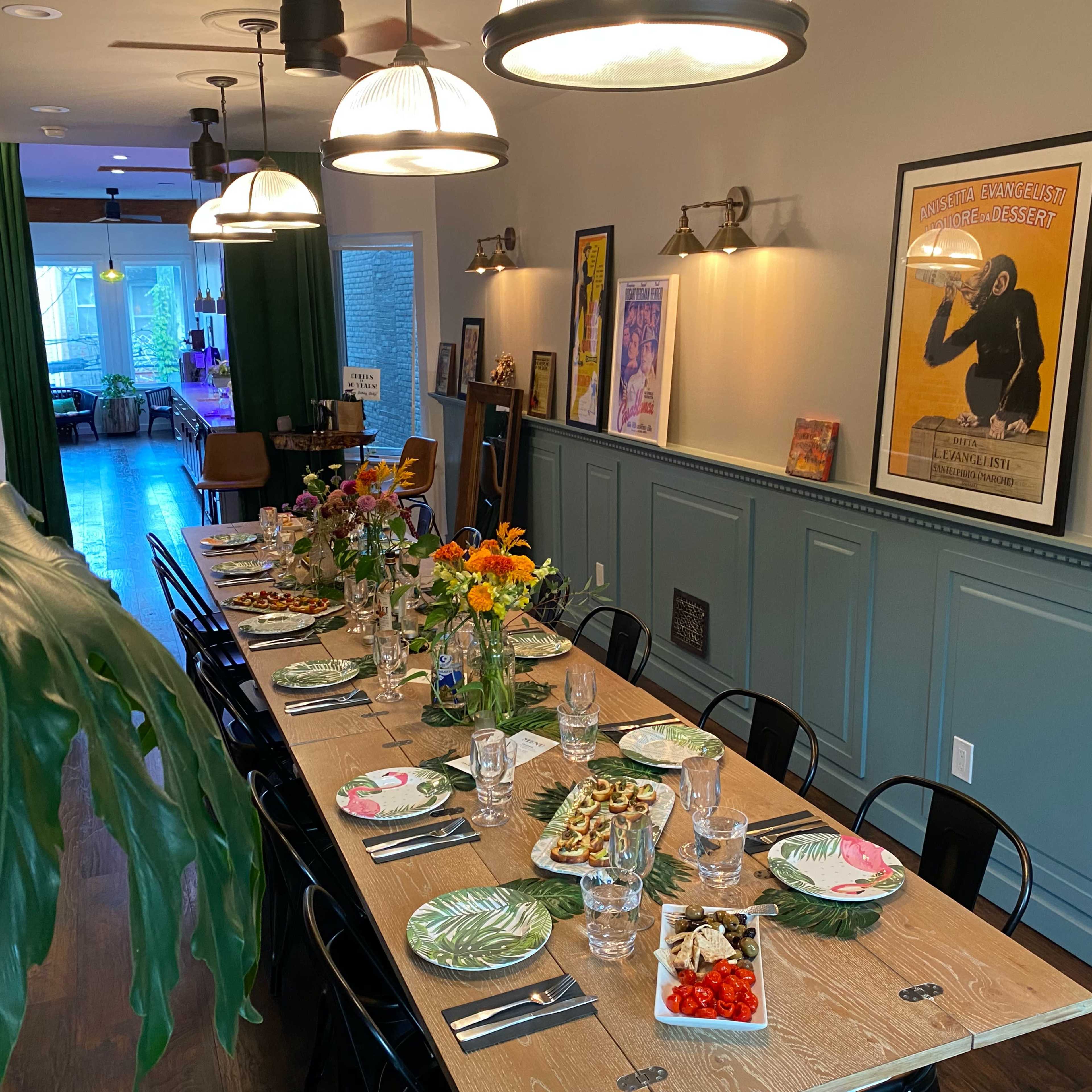 A long dining table with plates and glasses is set for a meal, surrounded by decorative plants and art on the walls in a well-lit room.