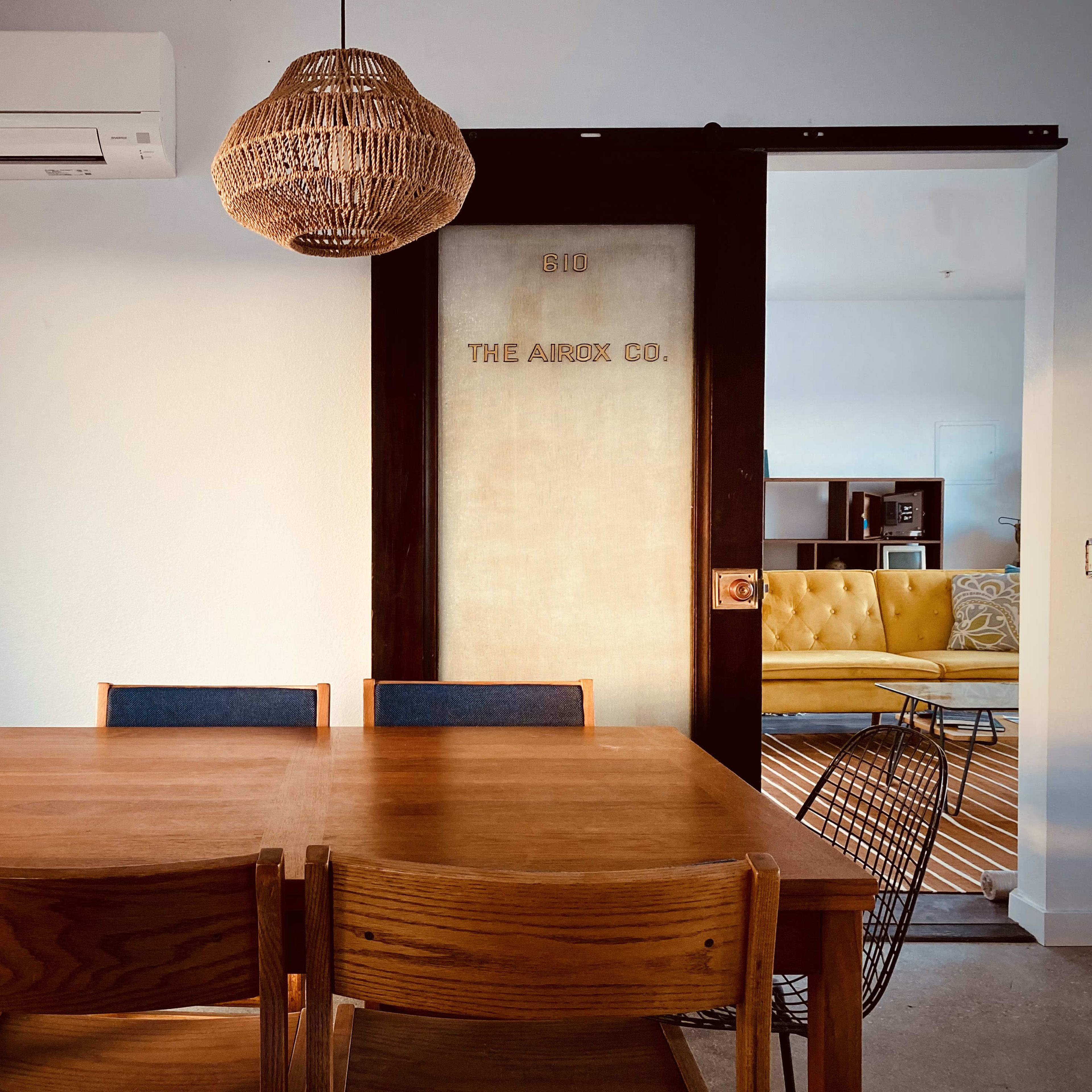 A wooden dining table with four chairs is positioned in front of a sliding door labeled "THE ARROW CO.", with a woven pendant light hanging above.