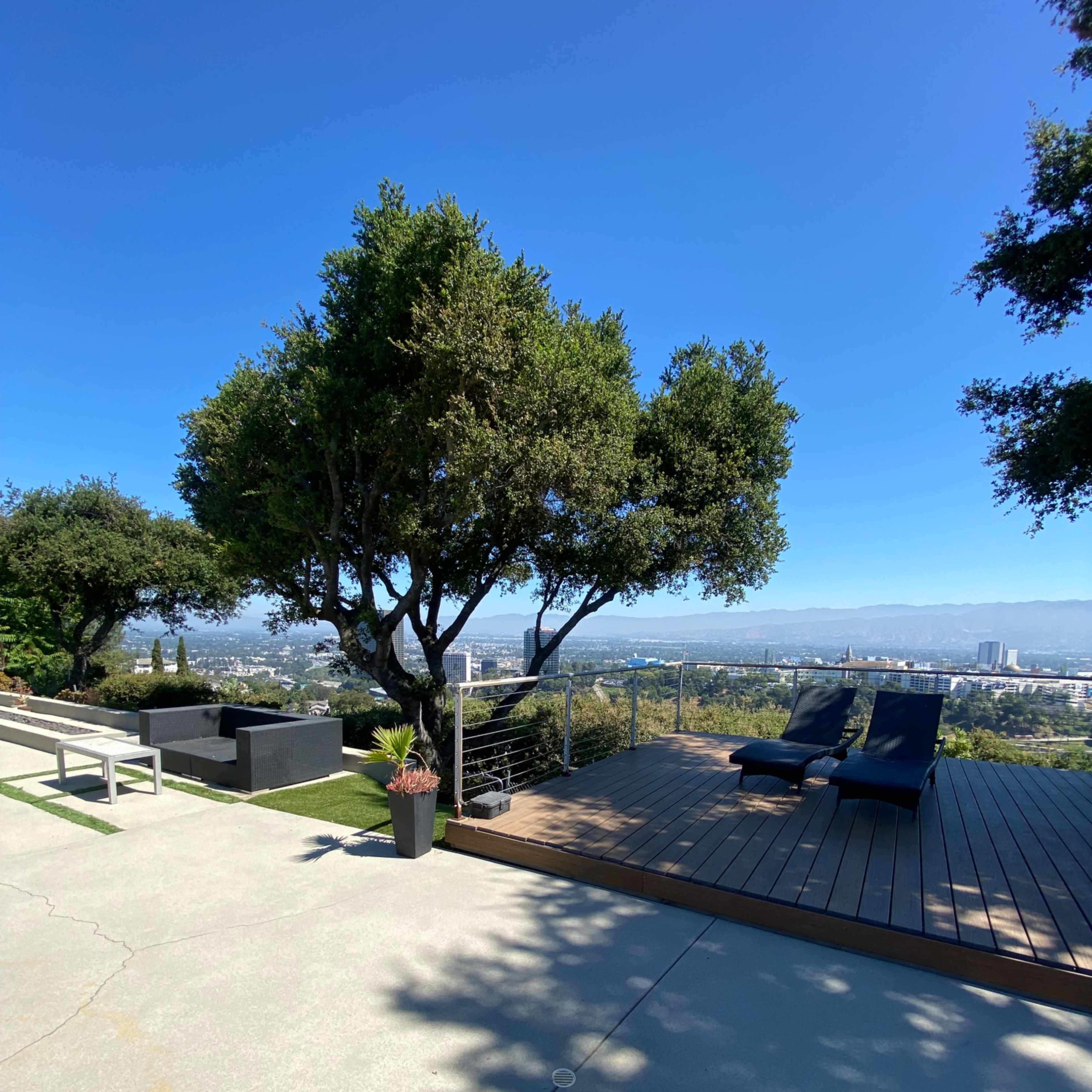 A clean outdoor space featuring a wooden deck, lounge chairs, a landscaped area with a planter, and a view of a cityscape under clear blue skies.