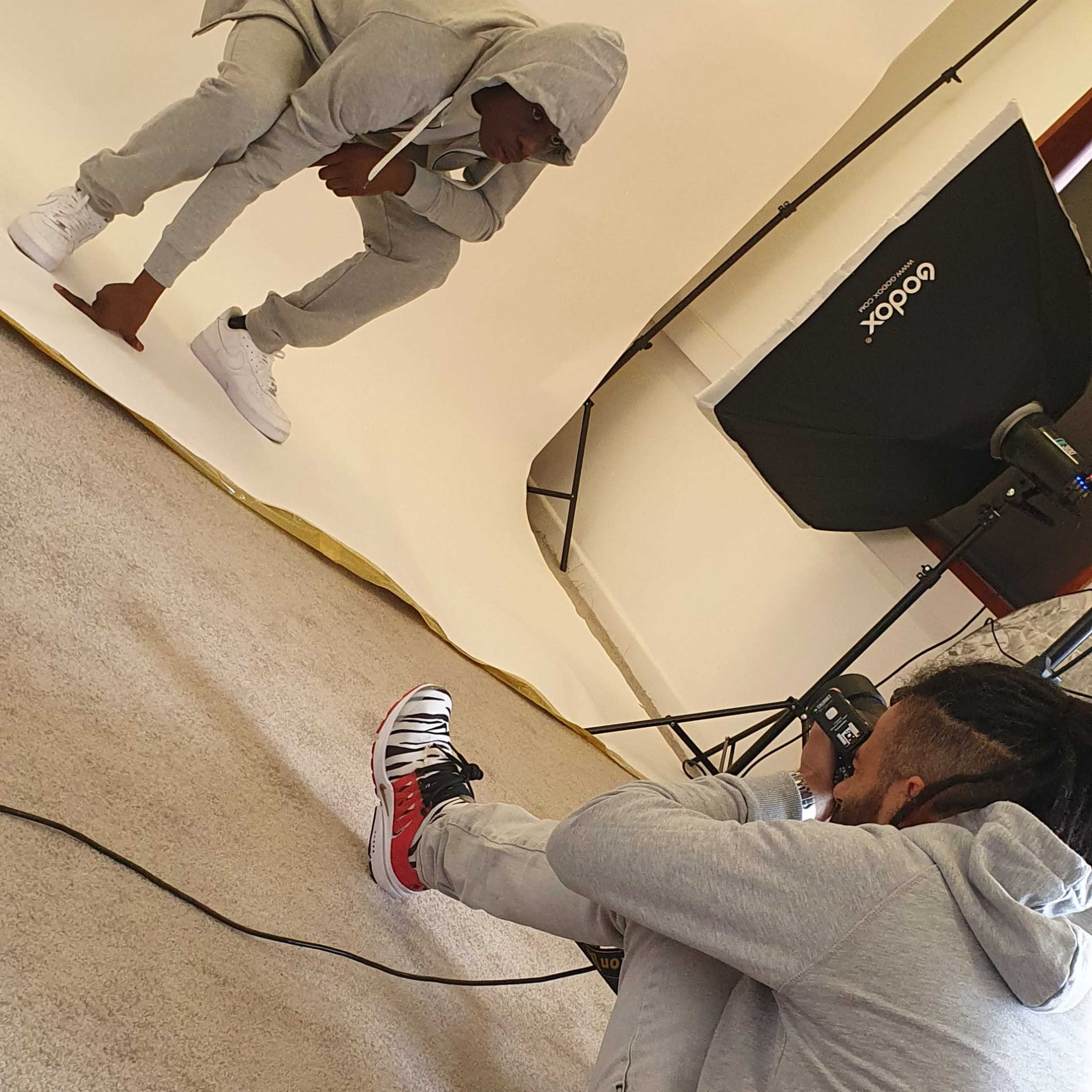 A photographer captures a seated model in a gray tracksuit against a neutral backdrop in a studio setting.