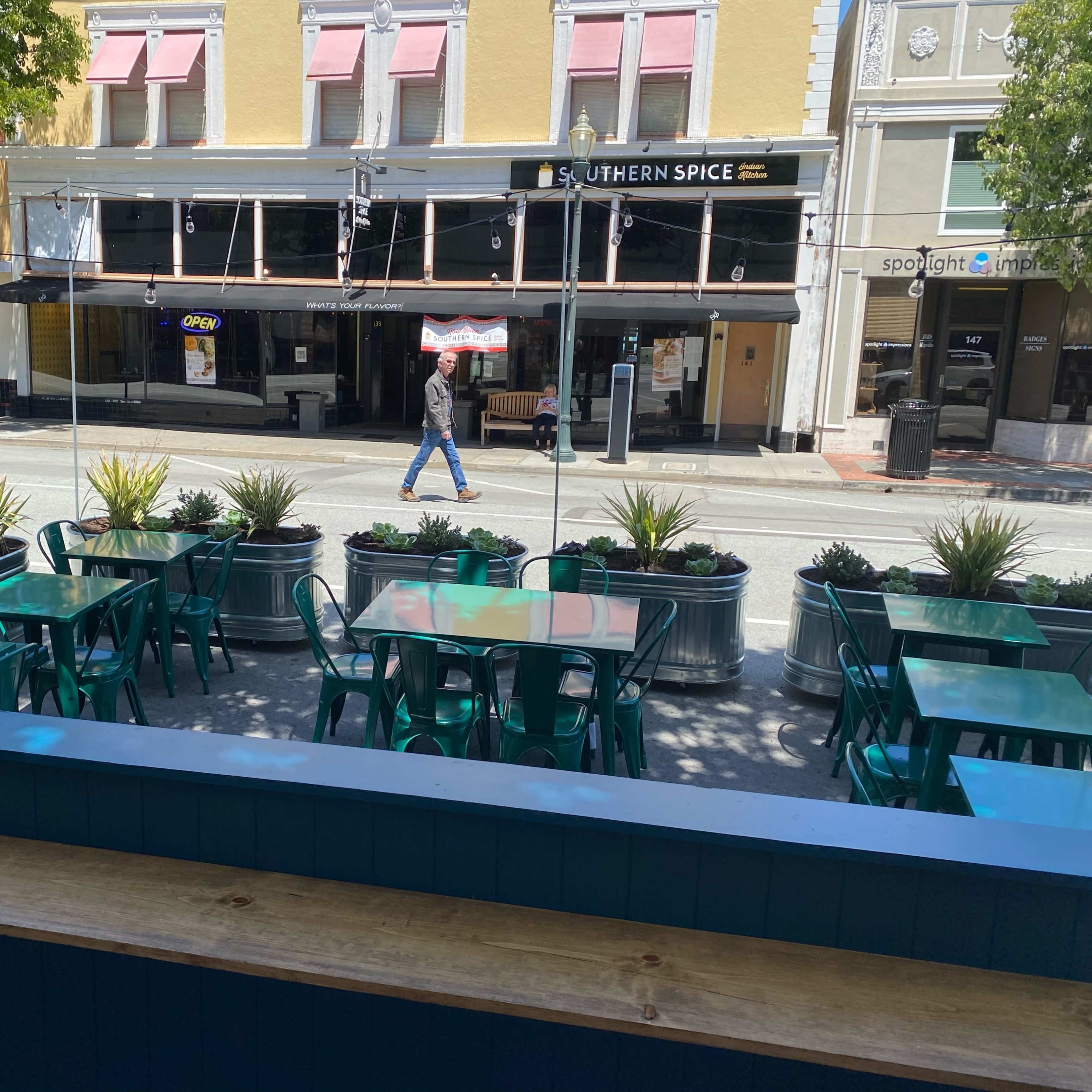A row of green tables and chairs are set outside a restaurant on a street lined with buildings.