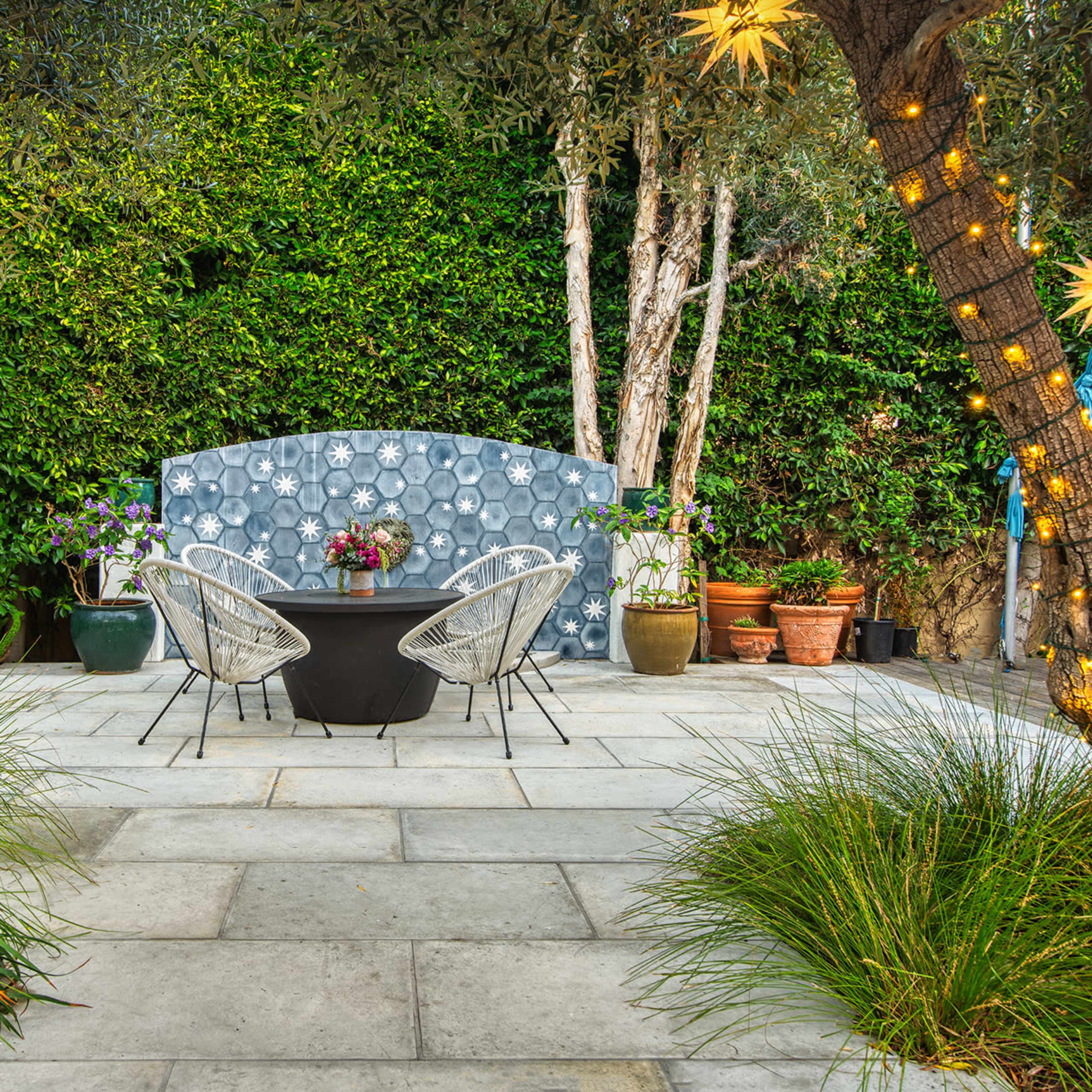 A stone patio features a round table with two chairs surrounded by lush greenery and decorated with string lights and star-shaped ornaments.