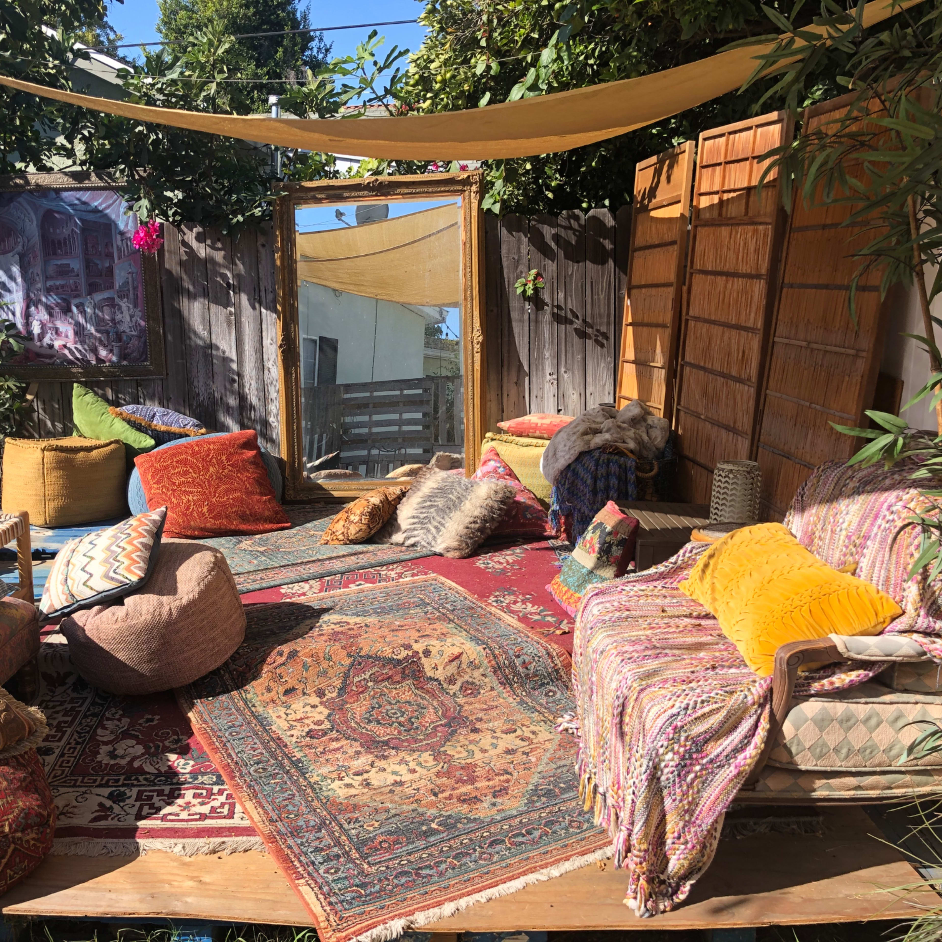 The scene features a cozy outdoor seating area adorned with various cushions, rugs, and a large mirror surrounded by greenery.