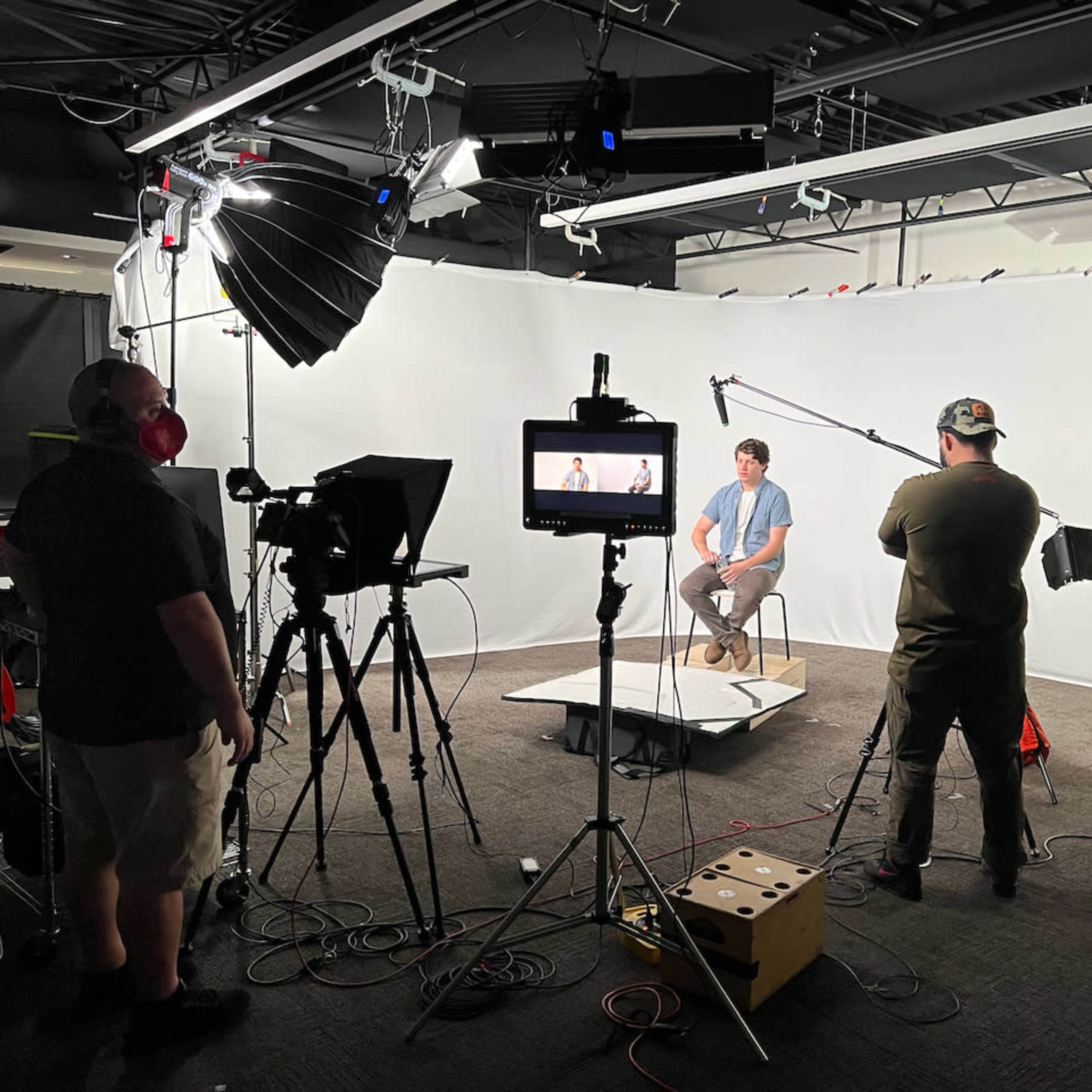 A small film crew sets up a video shoot in a studio with a subject sitting on a stool against a white backdrop.