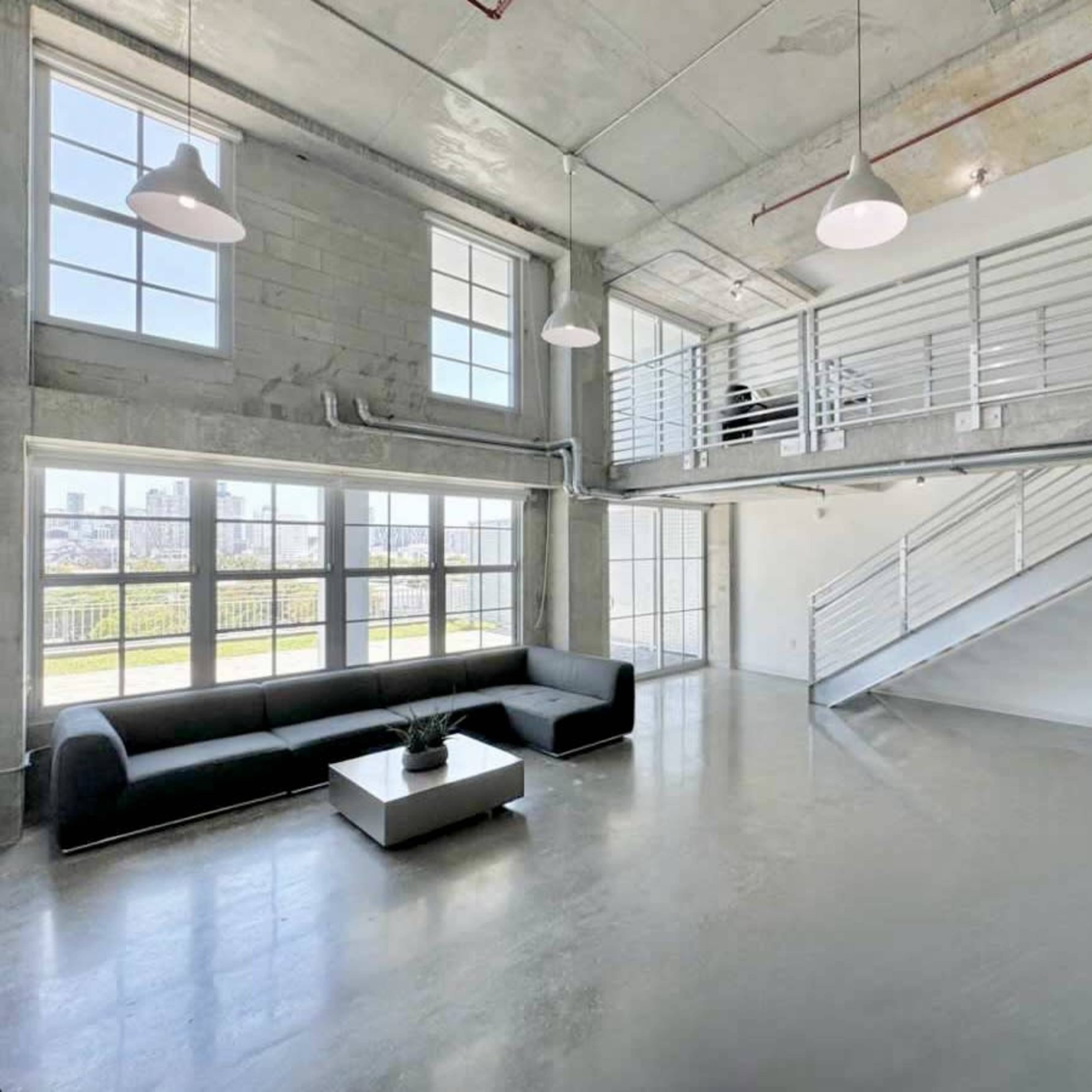 A modern, open-concept room with large windows, a gray sofa, a coffee table, and a staircase leading to an upper level.