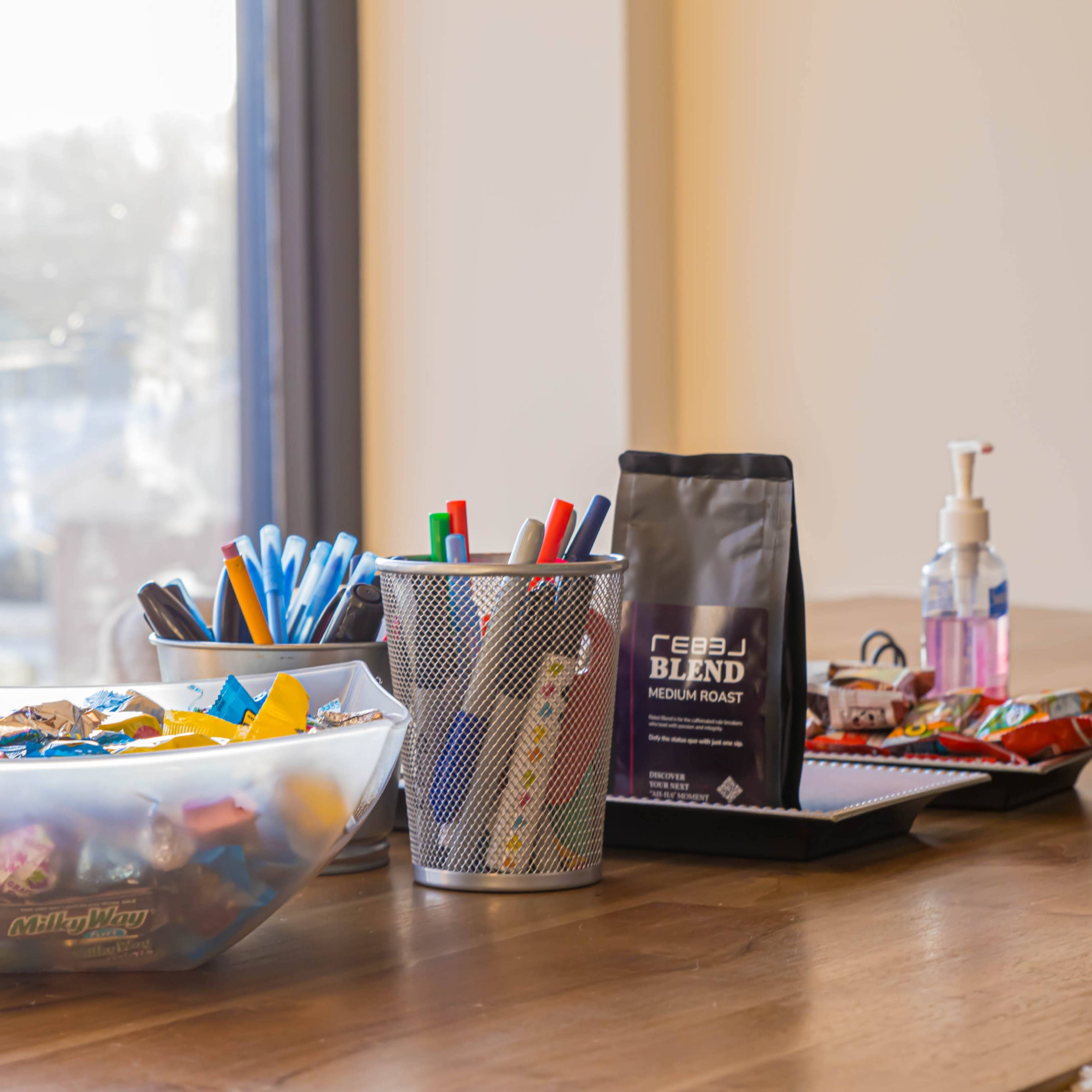 A wooden table displays a bowl of candy, a metal pencil holder with writing utensils, a coffee bag, a notepad, and a bottle of hand sanitizer.