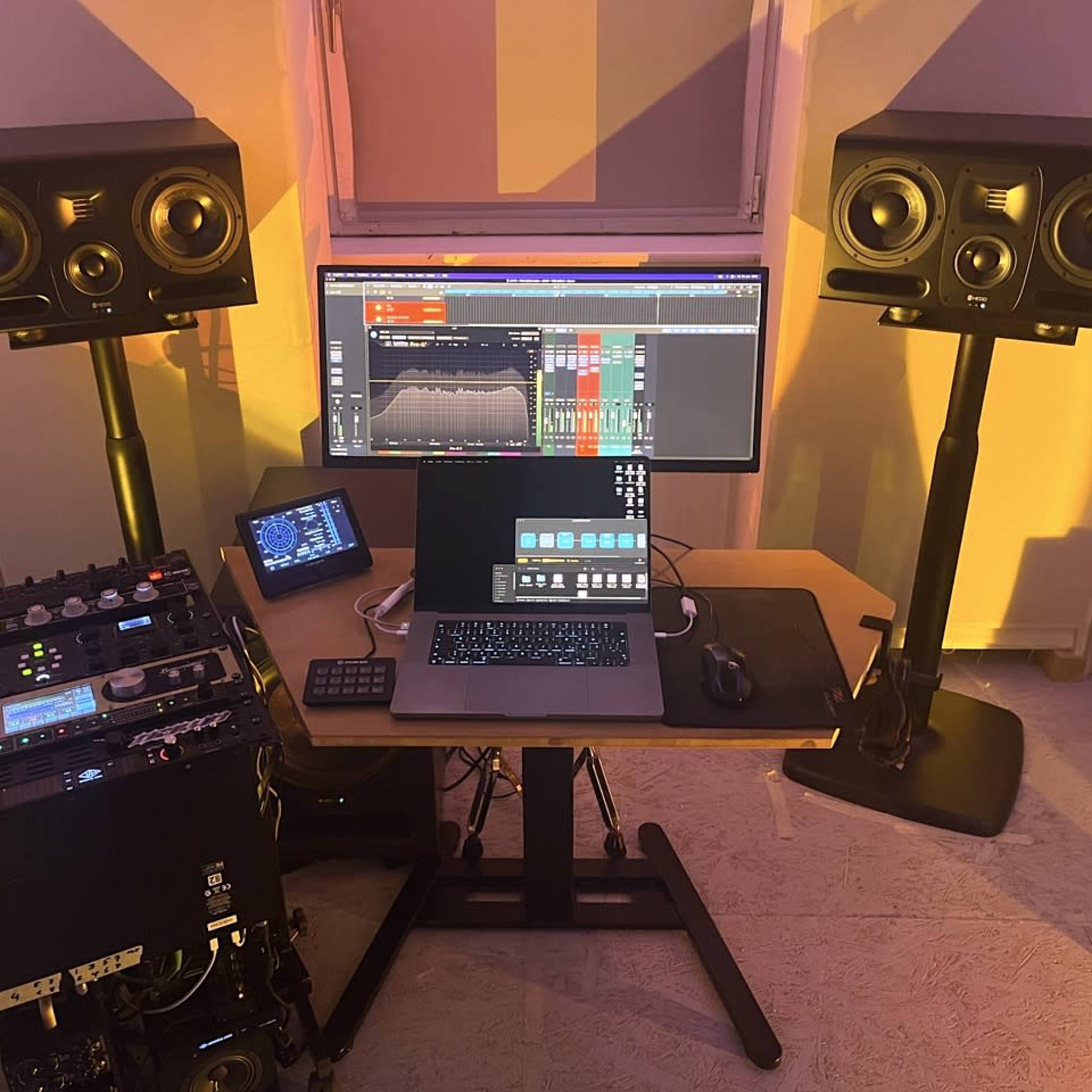A music production setup featuring a computer monitor, a laptop on a desk, and two studio monitors on stands.
