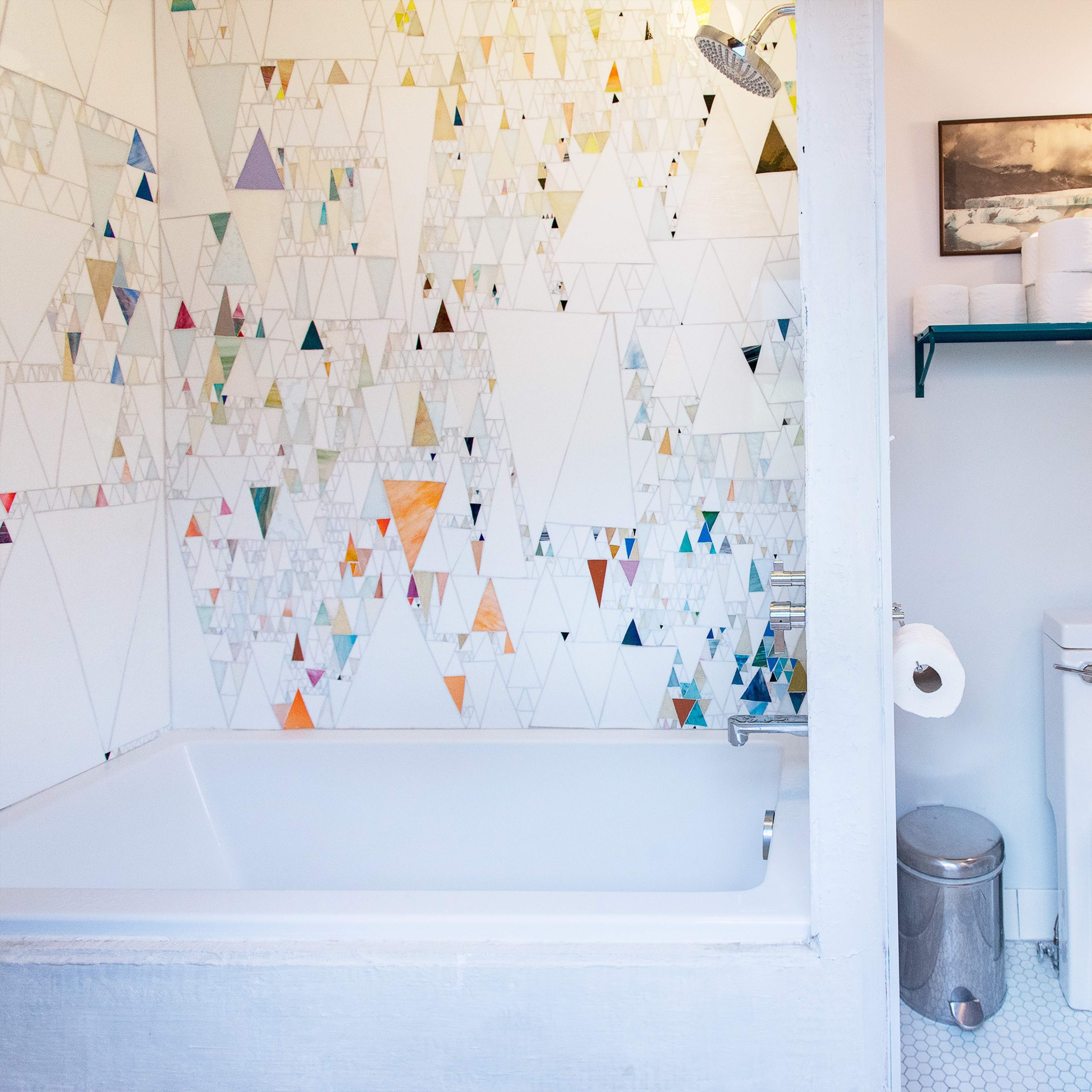A bathroom featuring a bathtub with a colorful triangular tile wall, a modern toilet, and a mirror above a shelf with bathroom essentials.