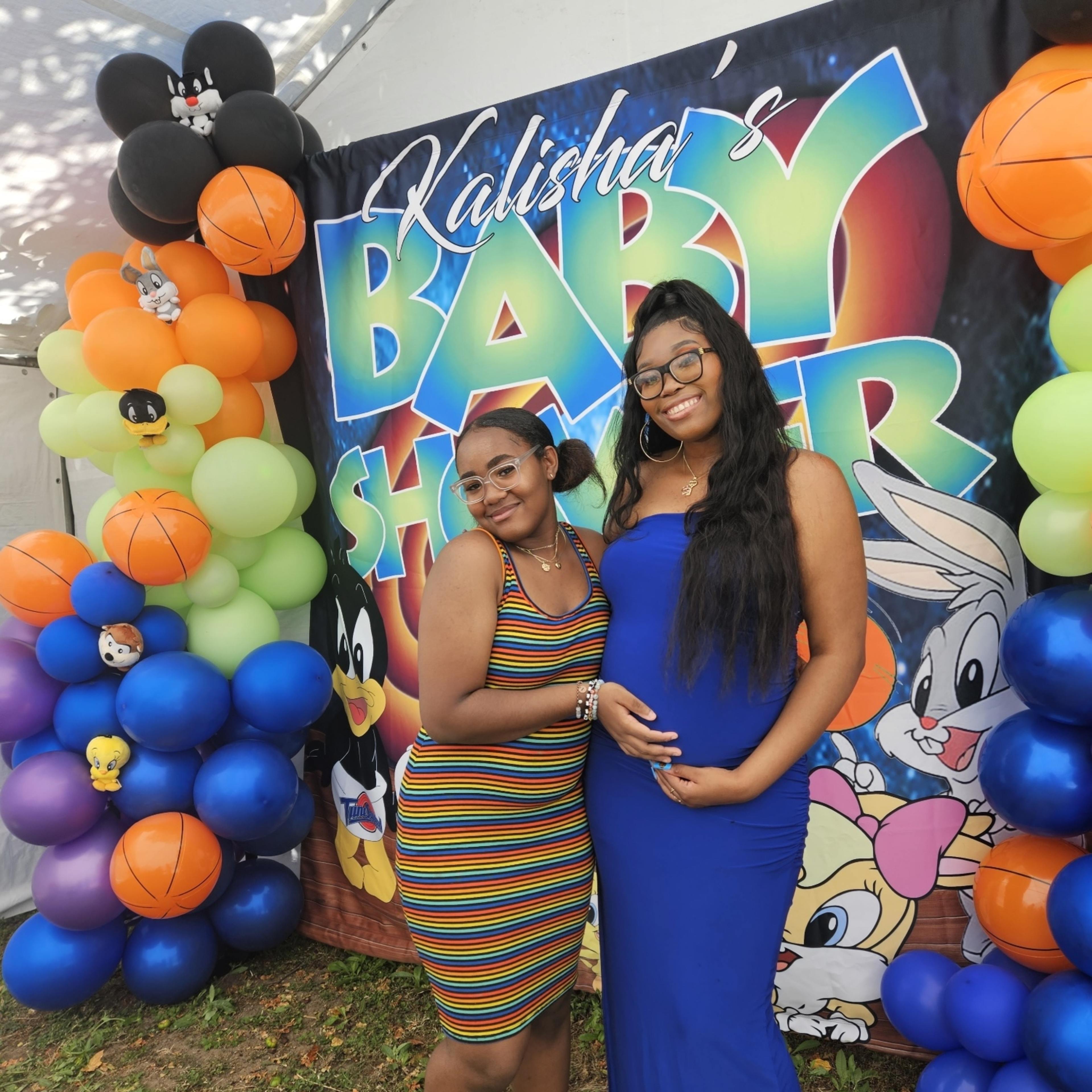 Two women pose in front of a colorful "Kalisha's Baby Shower" banner surrounded by balloons and cartoon characters.