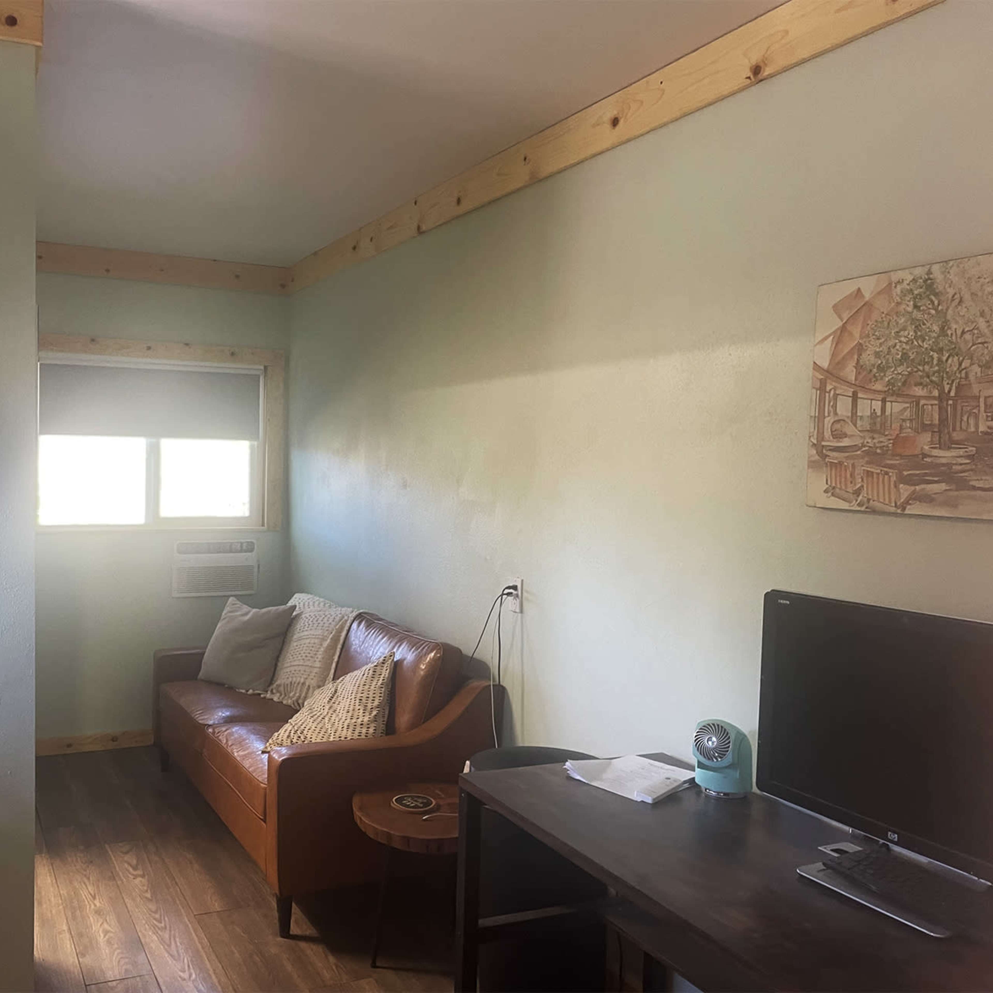 A room with a brown leather couch, a wooden table, a clock on the wall, and a window with an air conditioning unit.