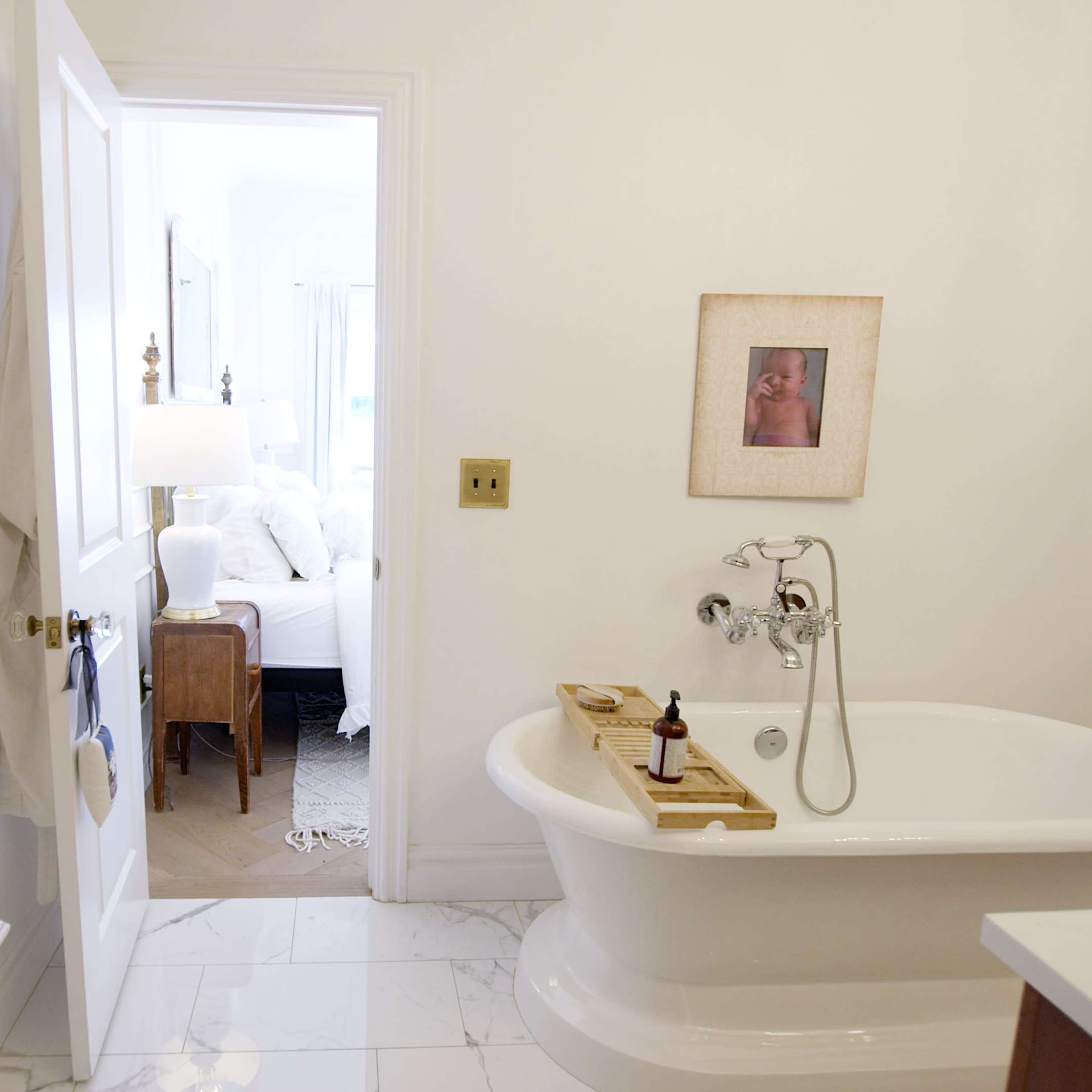 A bright bathroom features a freestanding bathtub, a doorway leading to a bedroom, and a few decorative items on a side table.