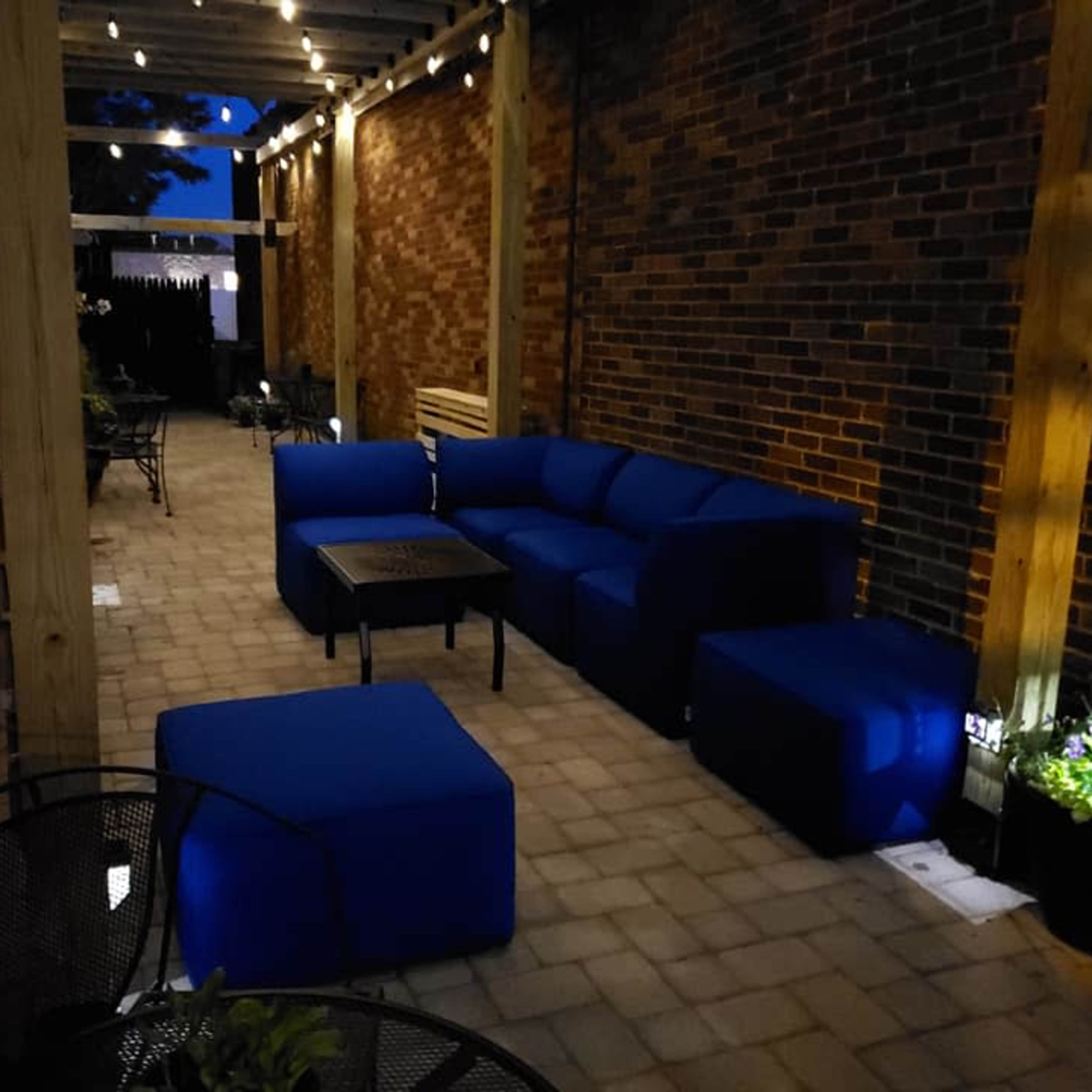 A cozy outdoor seating area featuring blue furniture arranged around a central table, illuminated by string lights and surrounded by brick walls.