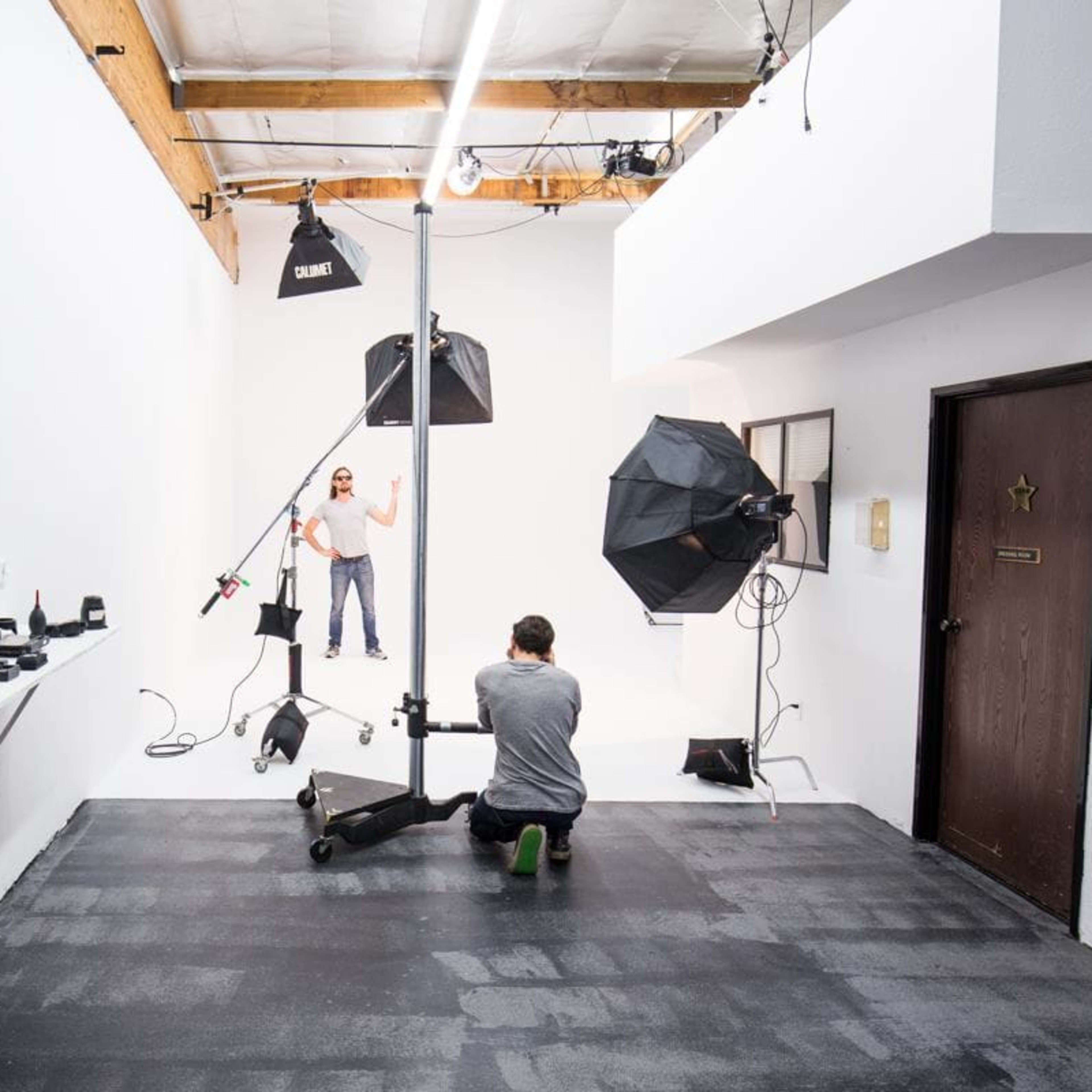 A photographer is capturing a model's portrait in a well-lit studio, equipped with various cameras and lenses displayed on a shelf to the left.