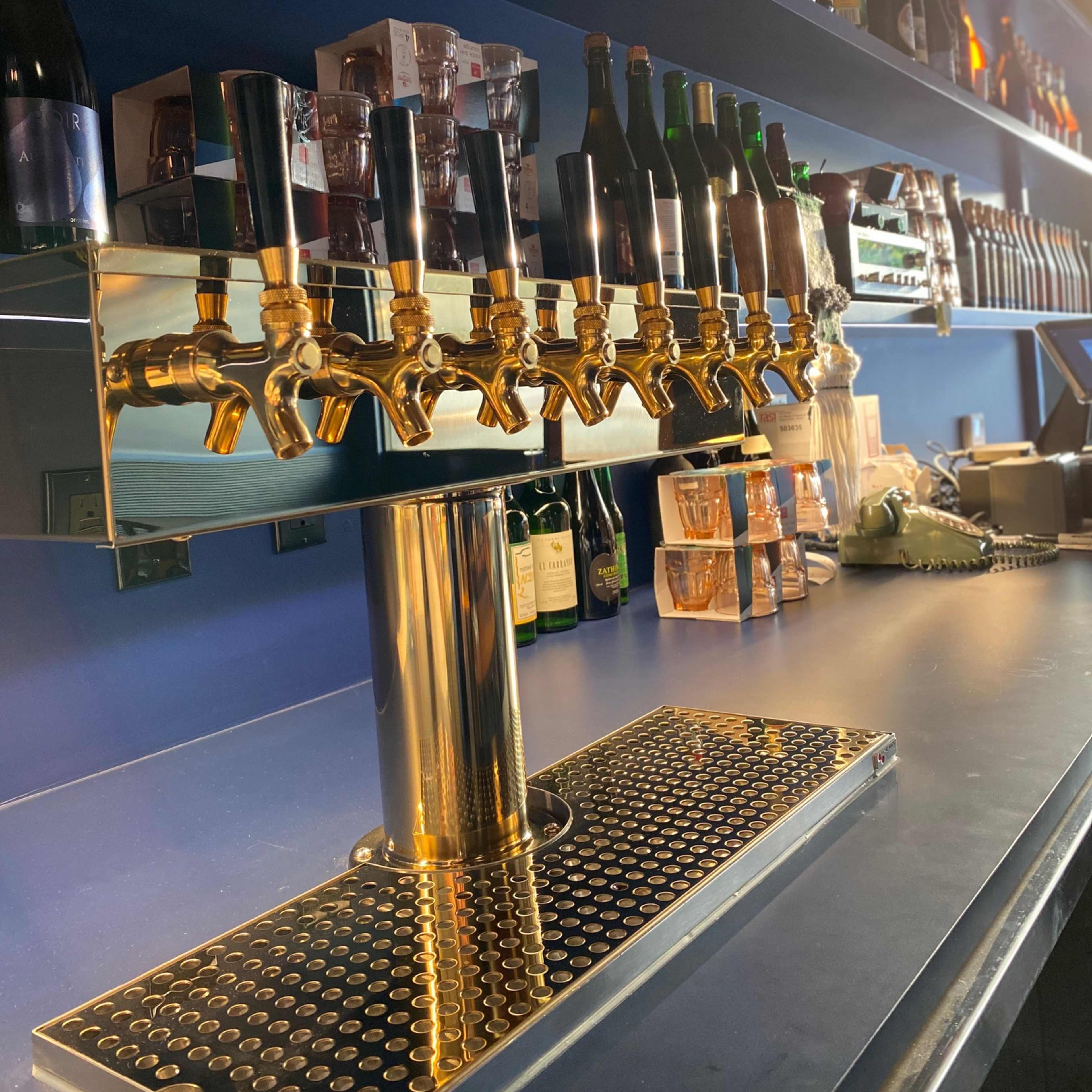 A polished bar with a row of eight shiny brass beer taps and shelves lined with various bottles in the background.
