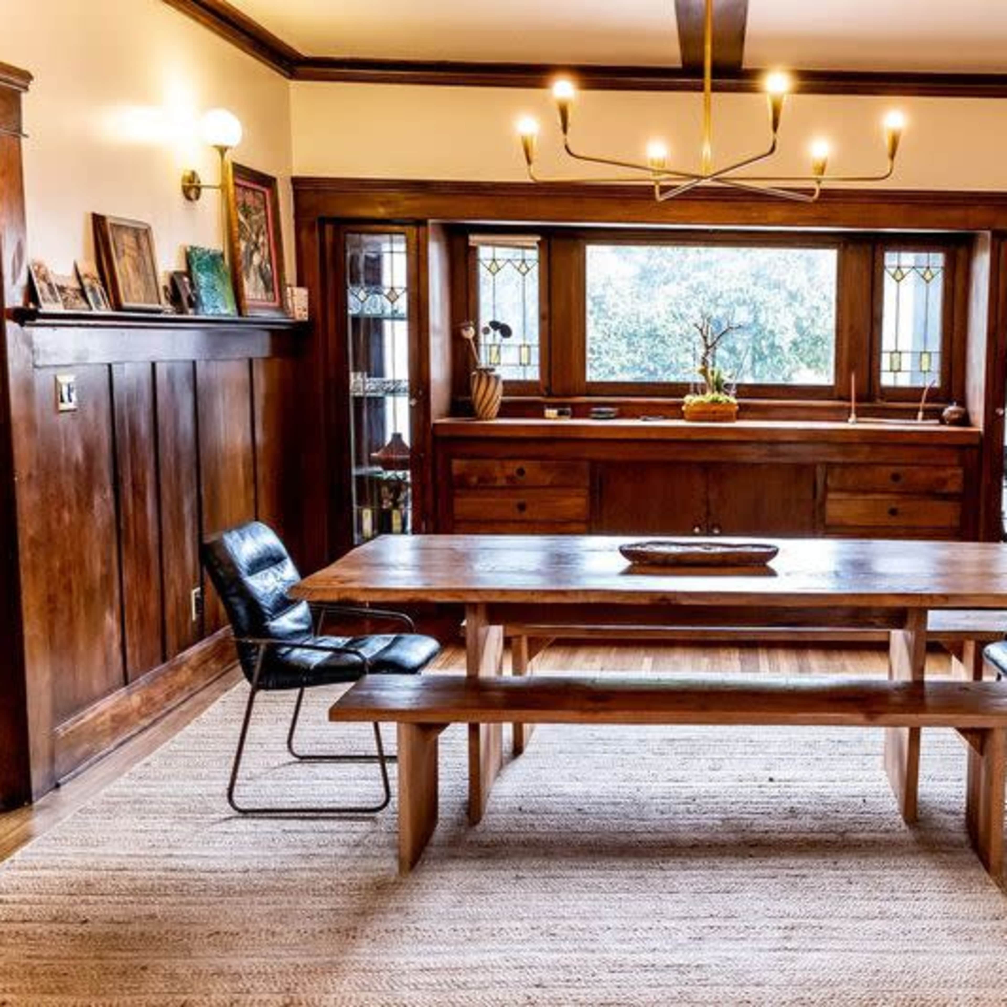A wooden dining table with a bench and two black chairs is positioned in a room with dark paneling and large windows.