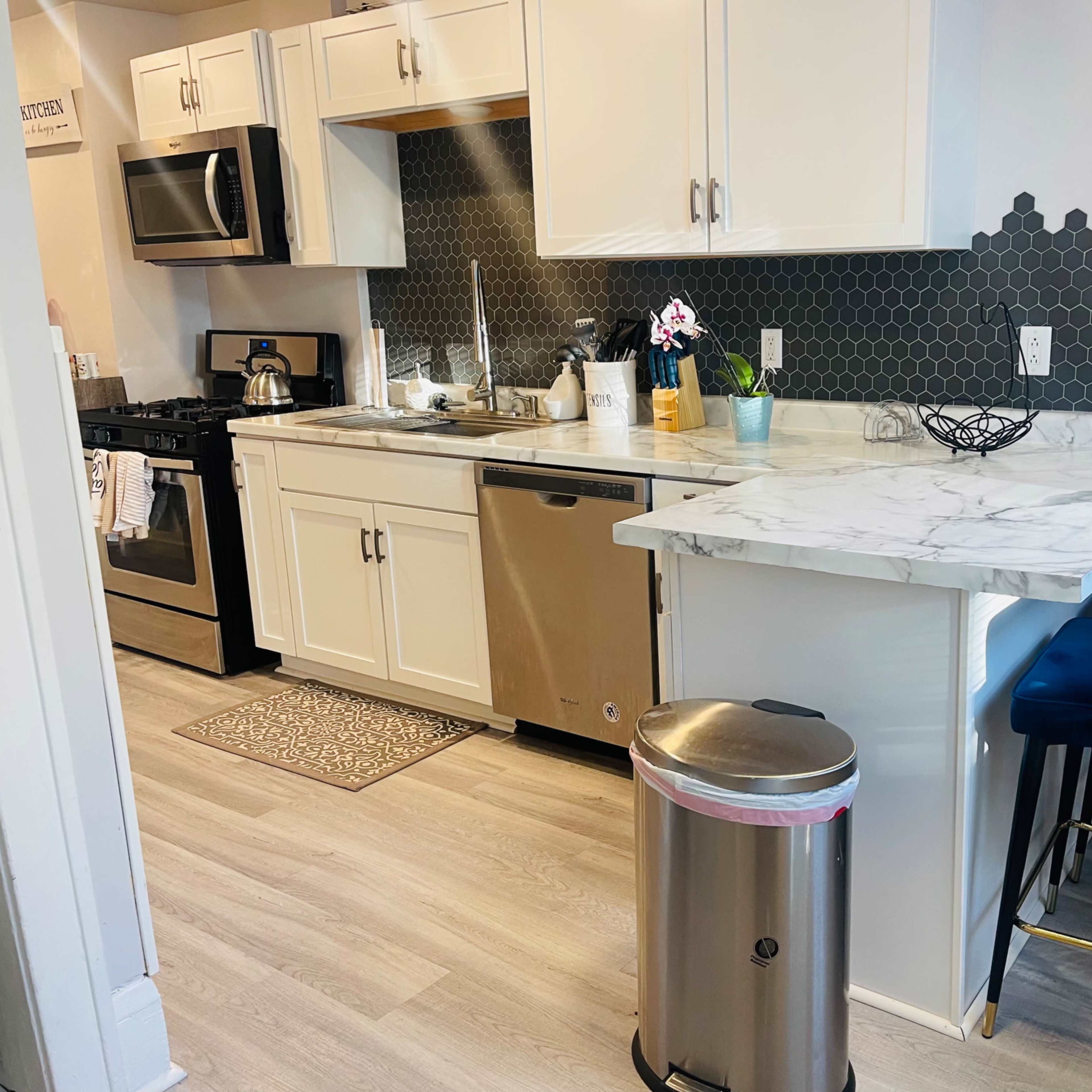 A modern kitchen features stainless steel appliances, white cabinets, a marble-topped island with blue bar stools, and a hexagonal backsplash.
