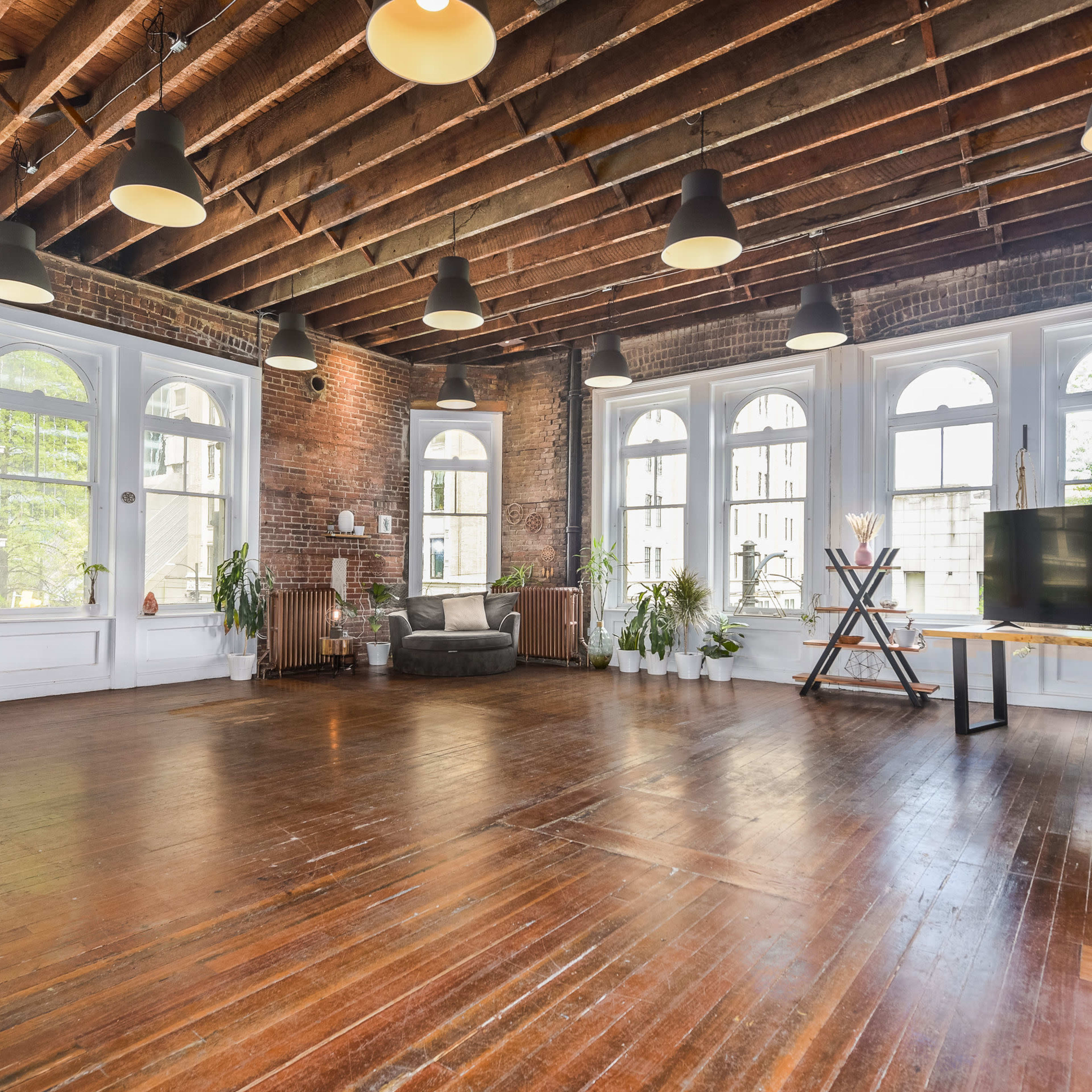 A spacious interior room with exposed wooden beams, large windows, and a mix of modern and rustic decor, featuring hardwood floors and potted plants.