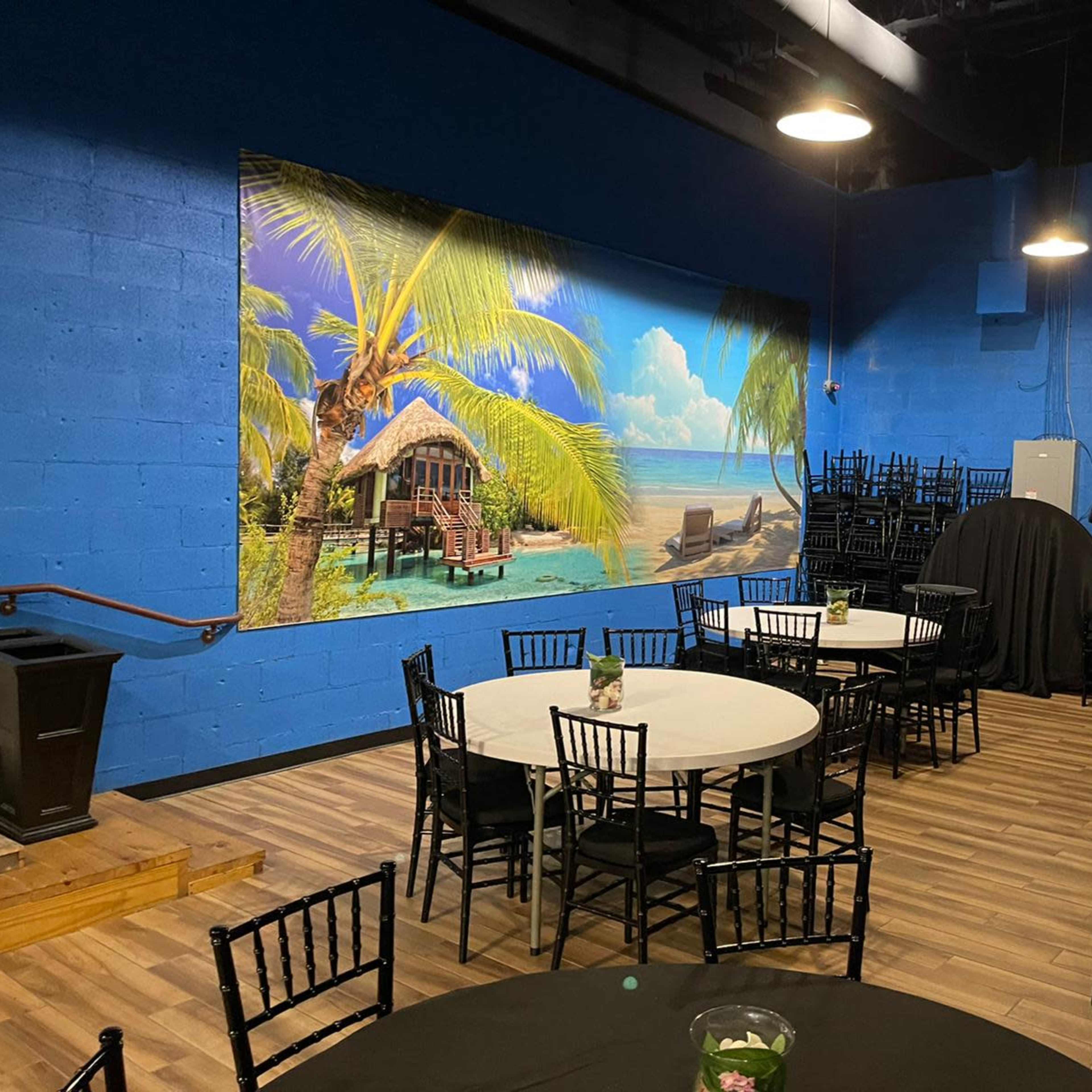 An event space with circular tables and black chairs, featuring a large tropical mural on a blue-painted wall.