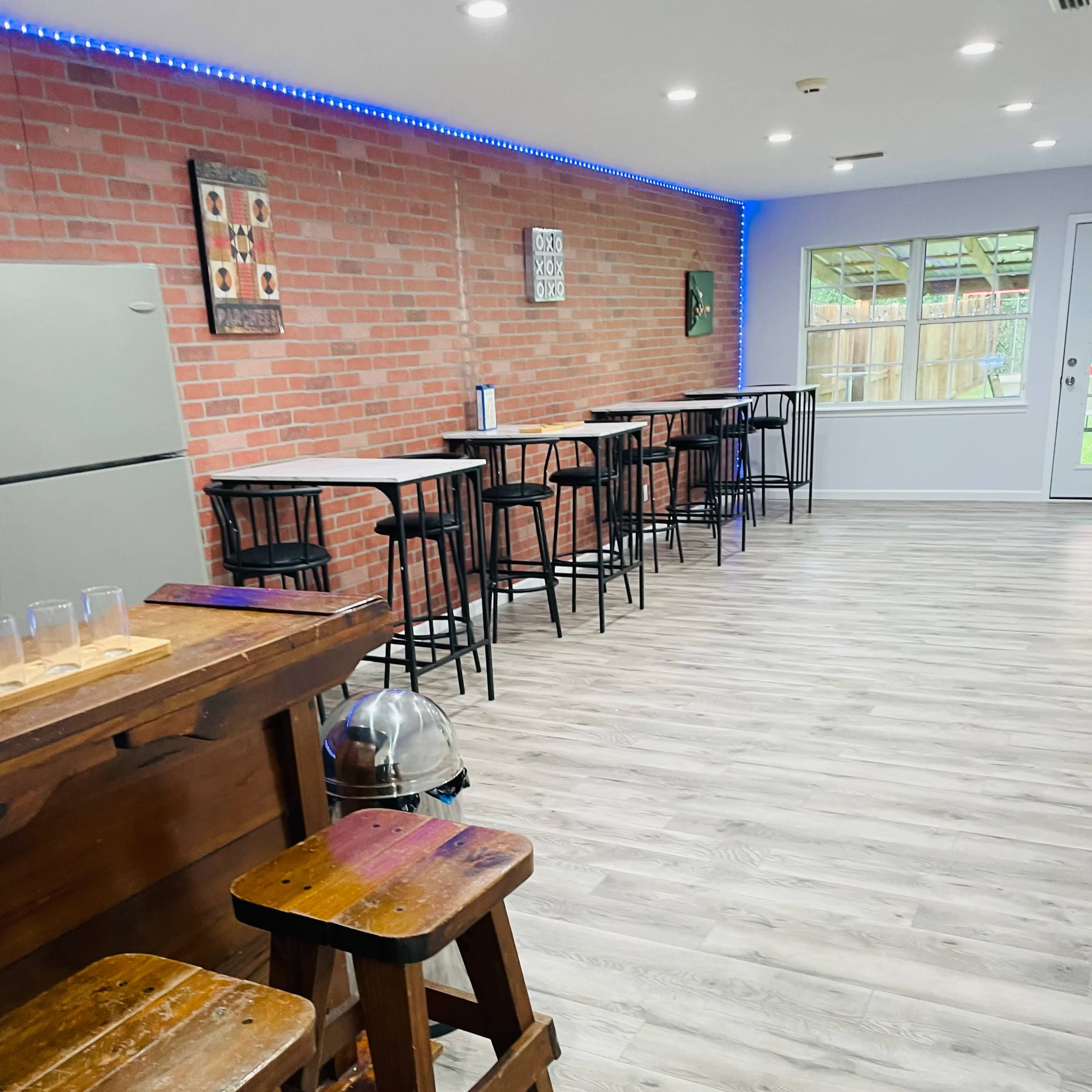 A room featuring a bar area with wooden stools, a refrigerator against a brick wall, and several high-top tables with chairs arranged along the opposite wall.