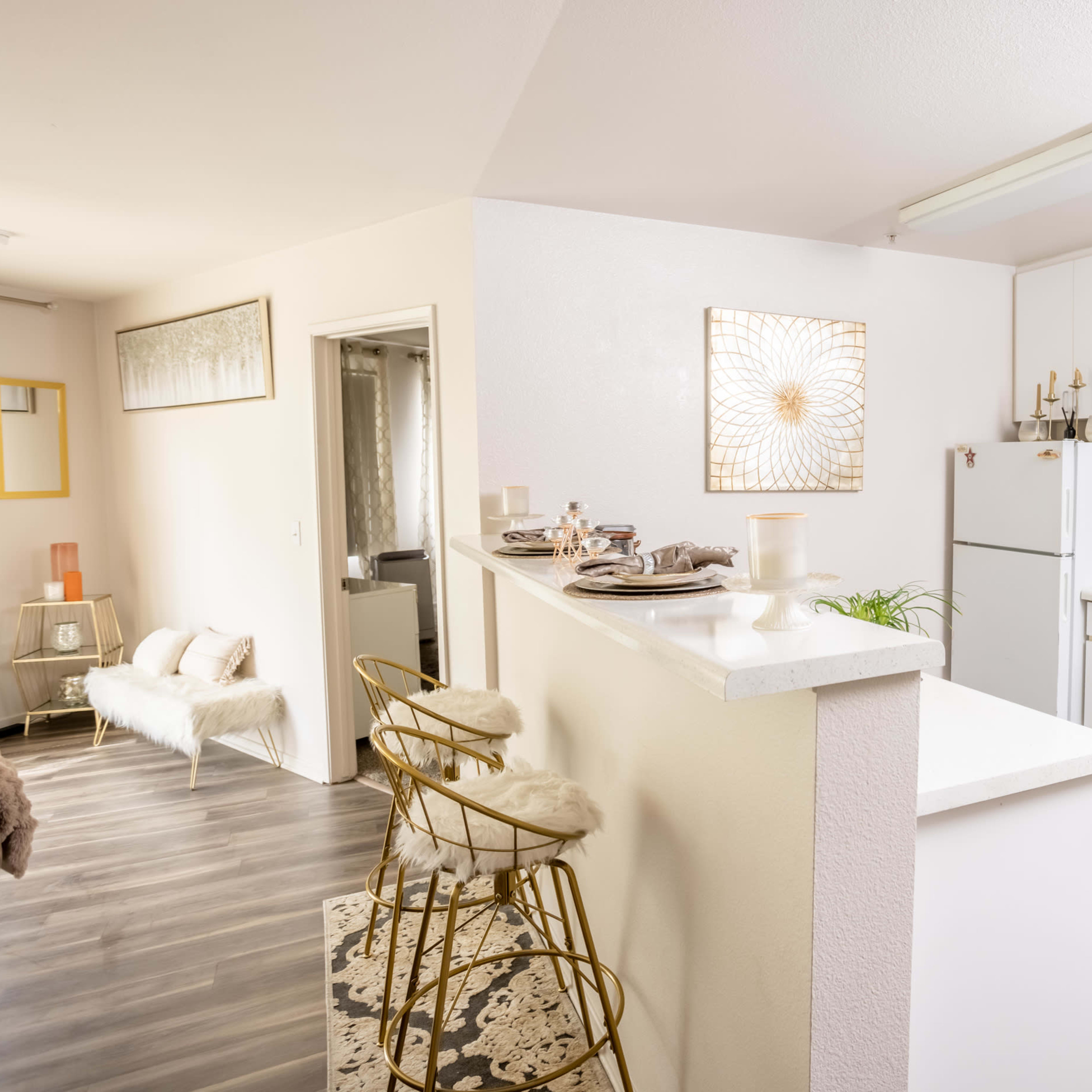 A modern kitchen features white cabinetry, an oven, a refrigerator, and bar seating with gold stools.