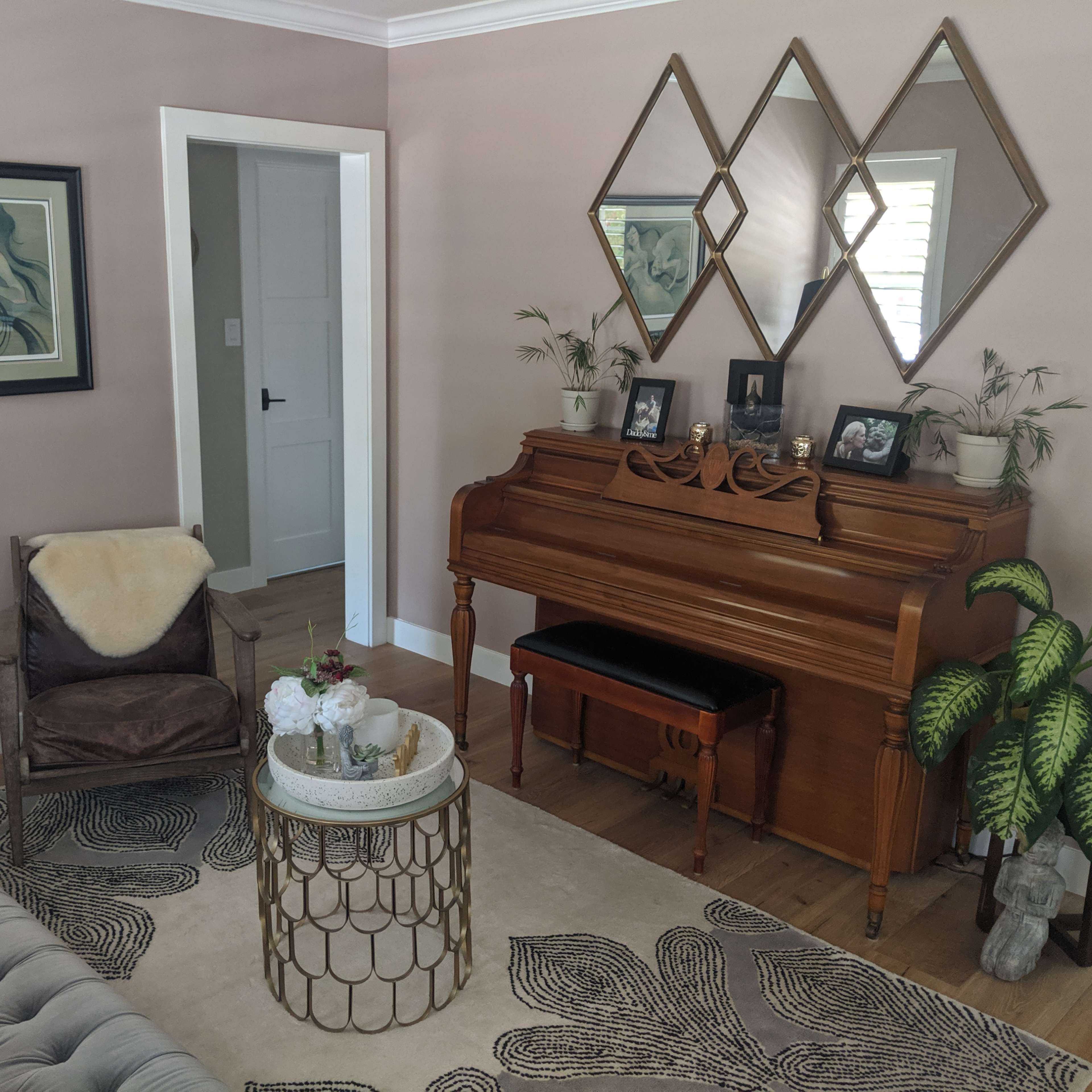 A well-furnished living room featuring a vintage piano, a round coffee table with flowers, a patterned area rug, and a large mirror with geometric shapes on the wall.