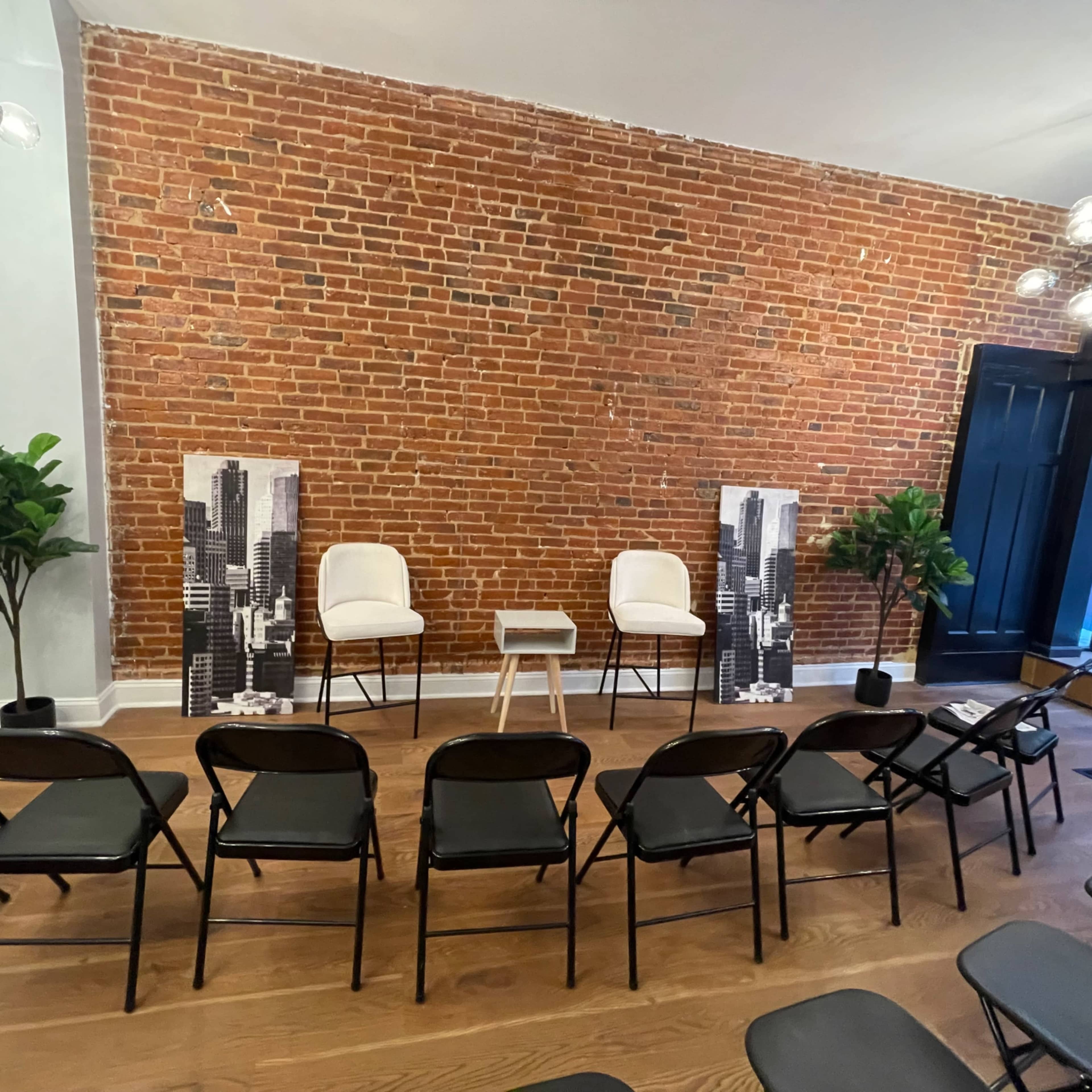 The scene features a room with exposed brick walls, two chairs facing a small table, and rows of folding chairs arranged in front of them, flanked by potted plants.