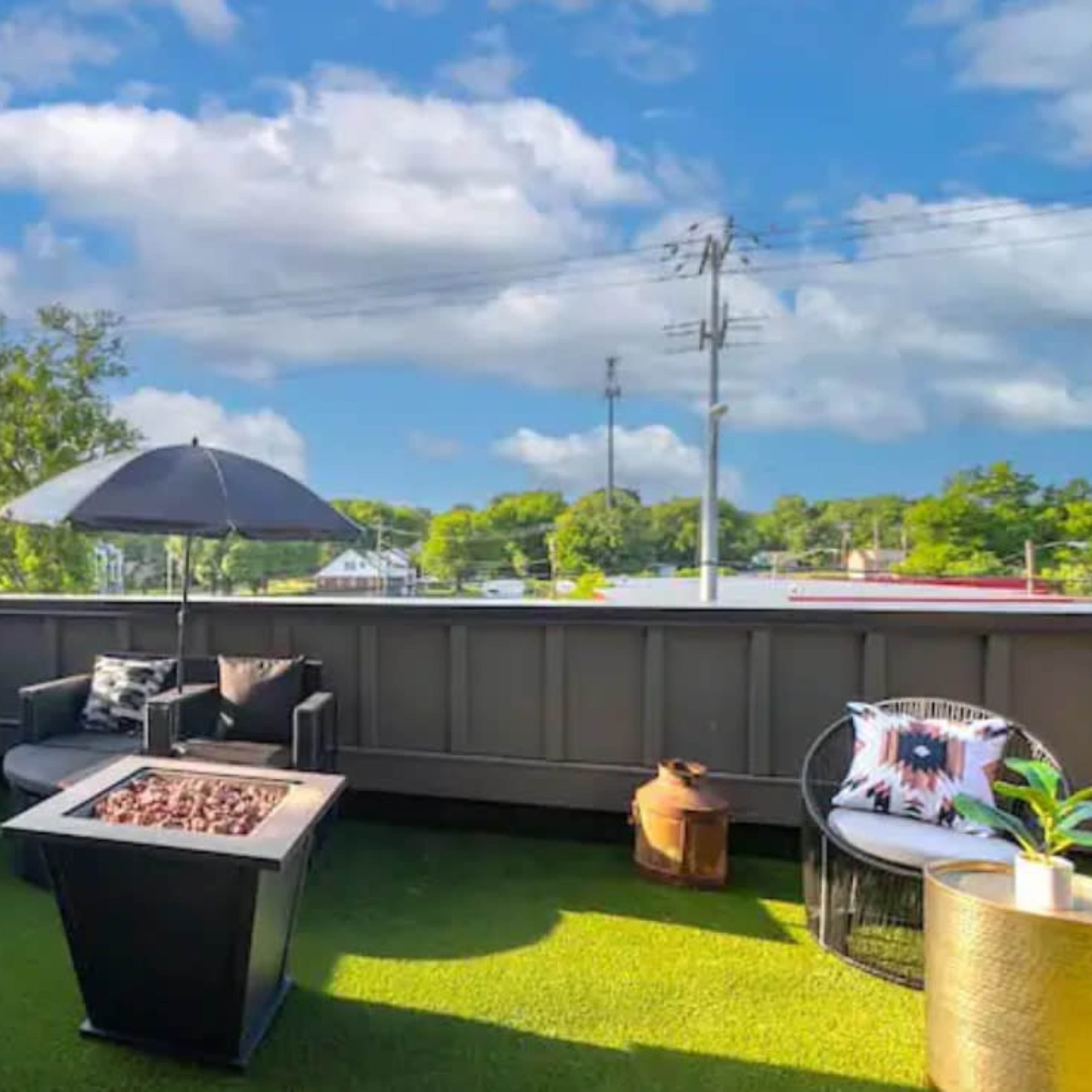 A rooftop patio features a seating area with circular couches, a fire pit, and an umbrella, surrounded by greenery and utility poles in the background.