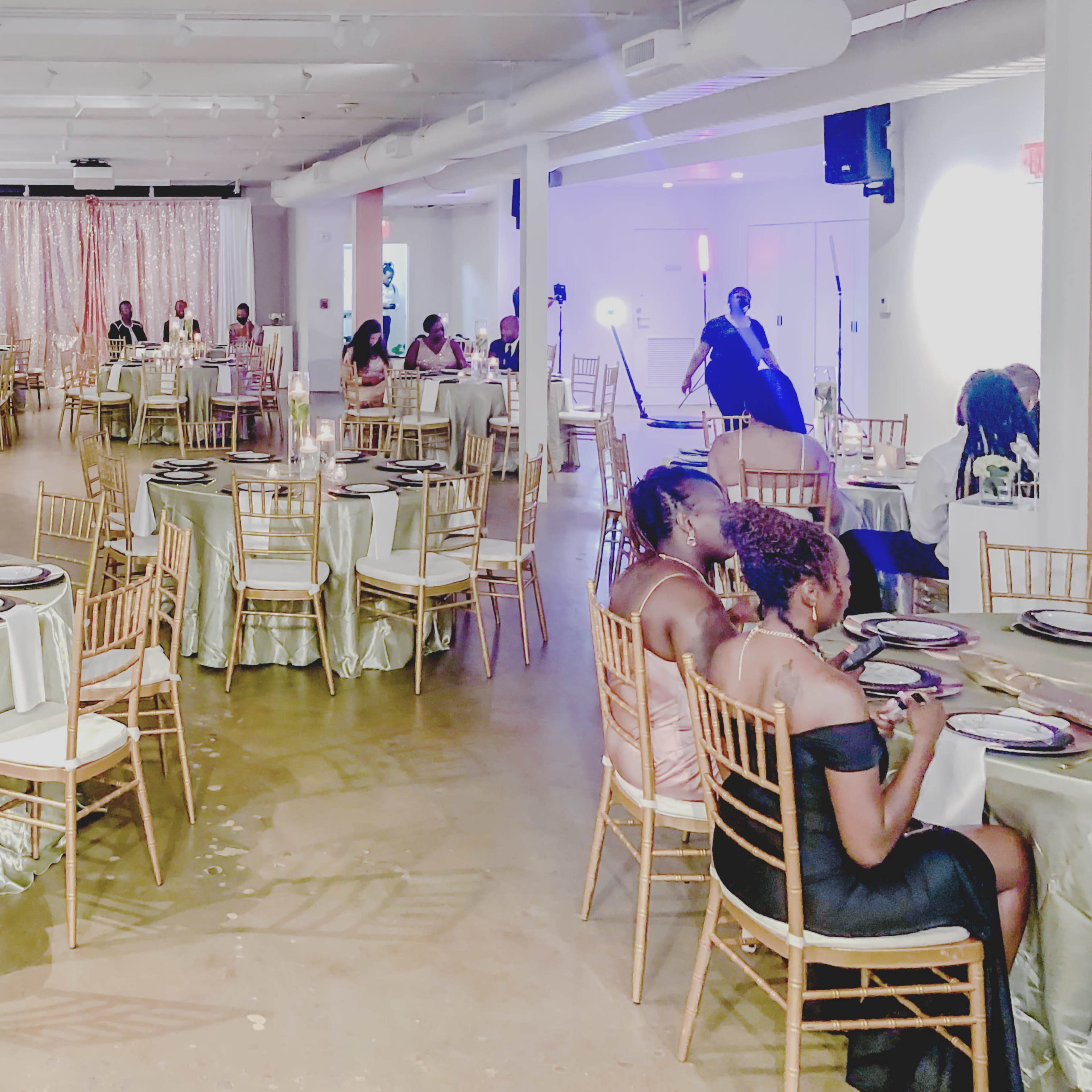 A spacious event venue is set up for a formal gathering, featuring elegantly dressed tables with gold chairs, decorative balloons, and a shimmering backdrop.