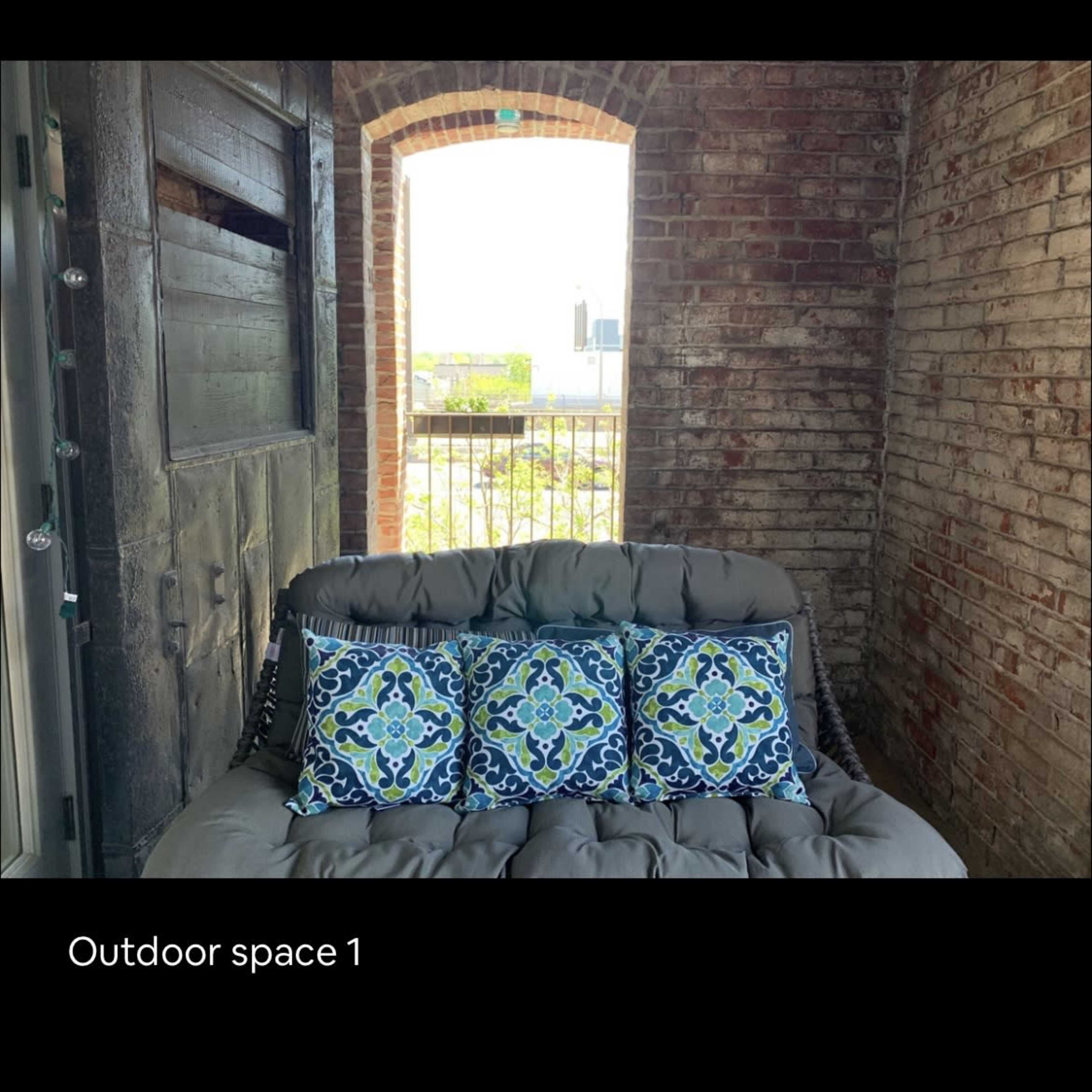 A cozy outdoor seating area with a patterned cushion sofa and a view through a window framed by brick walls.