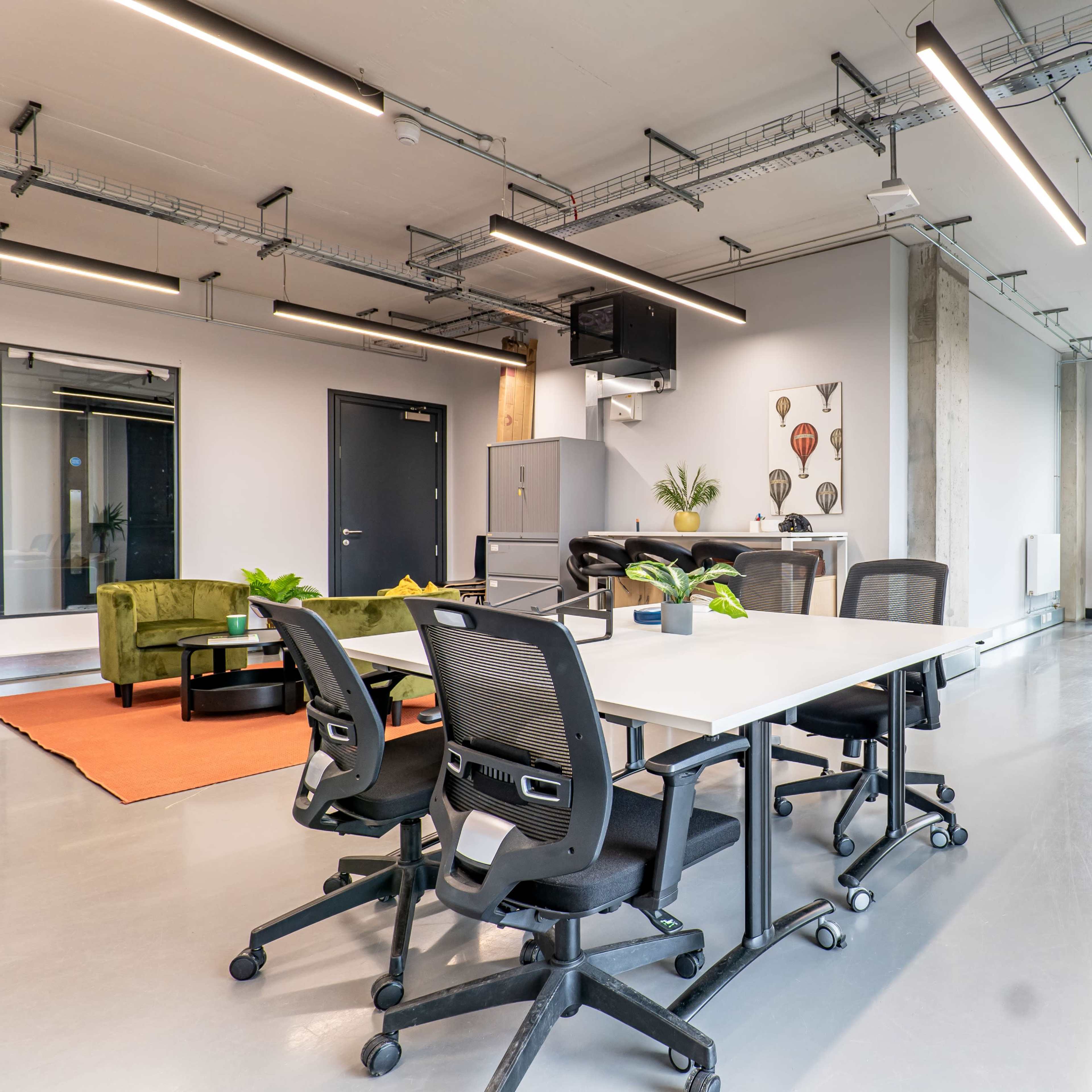 A modern office space featuring a large table with chairs, a green couch, and plants, with large windows providing natural light.